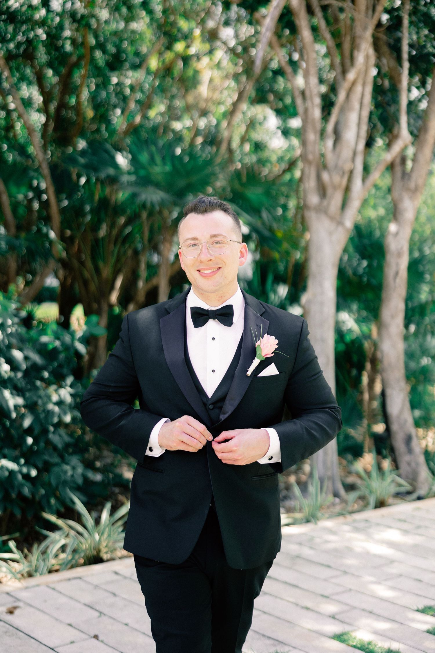Smiling groom in black tuxedo with bow tie and pink boutonniere, standing outdoors among lush green trees.