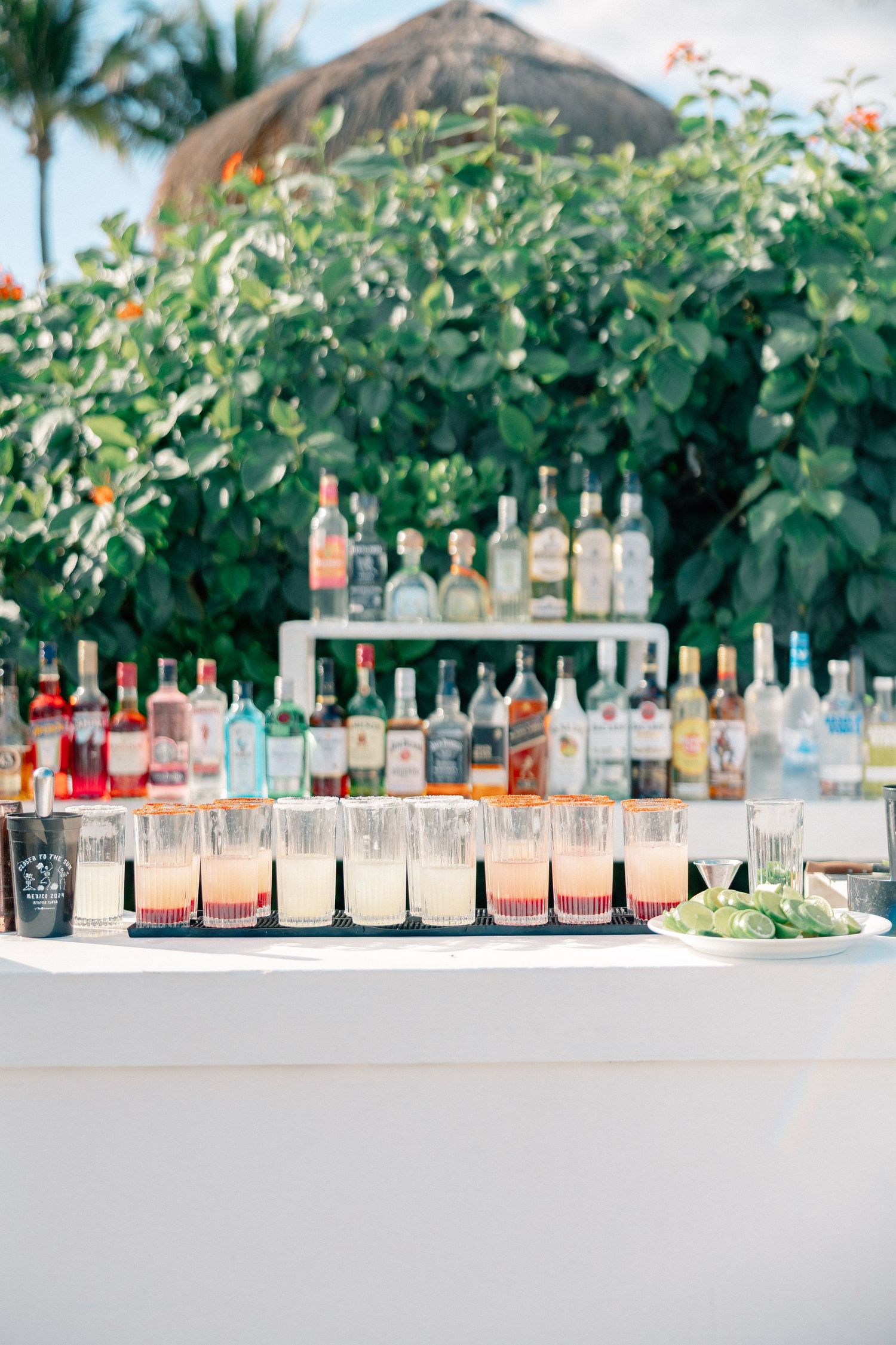 Outdoor bar display with colorful cocktails lined up and liquor bottles on shelves, surrounded by lush tropical greenery.