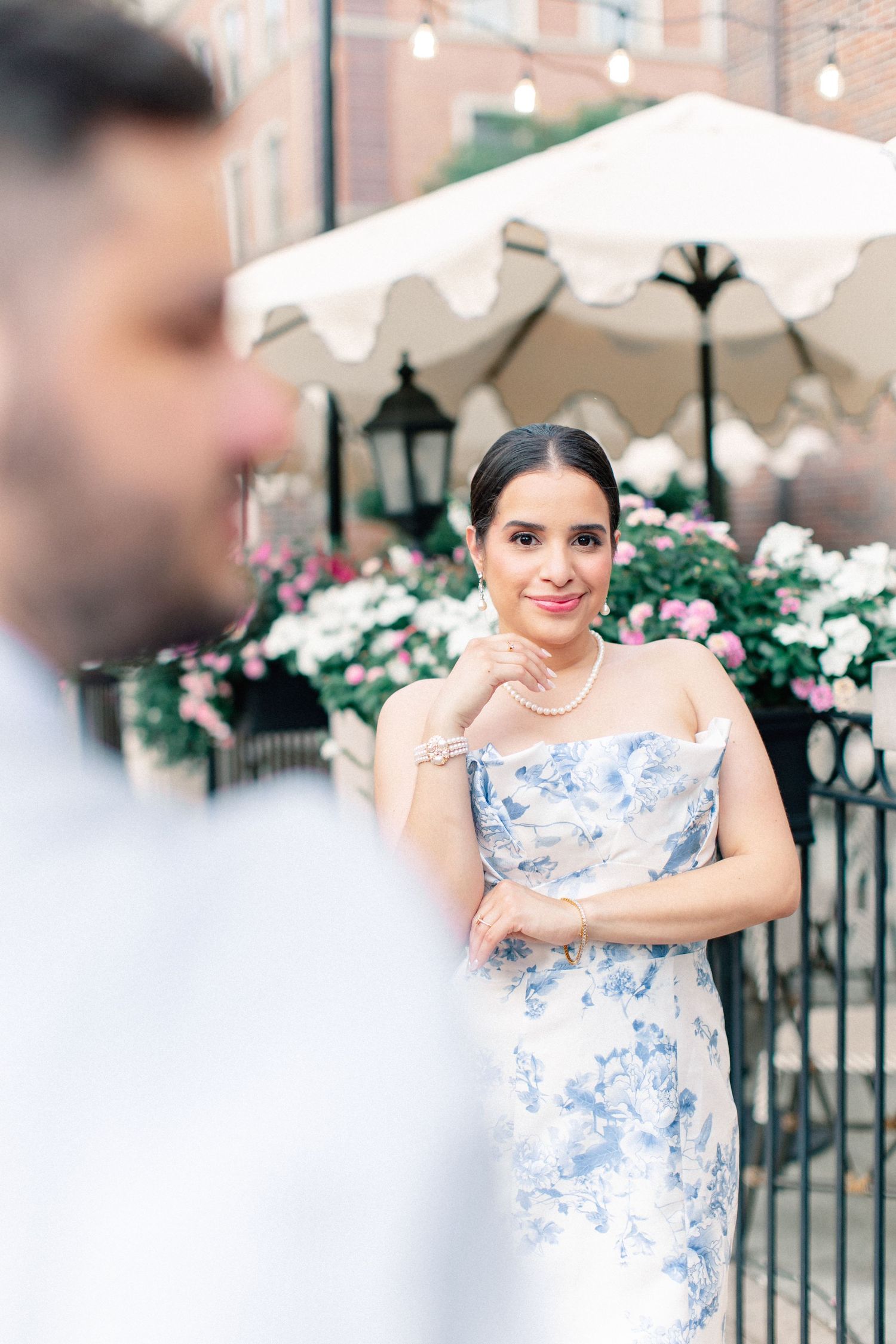 Bride in strapless blue floral dress with pearl necklace smiling at groom during outdoor engagement session.