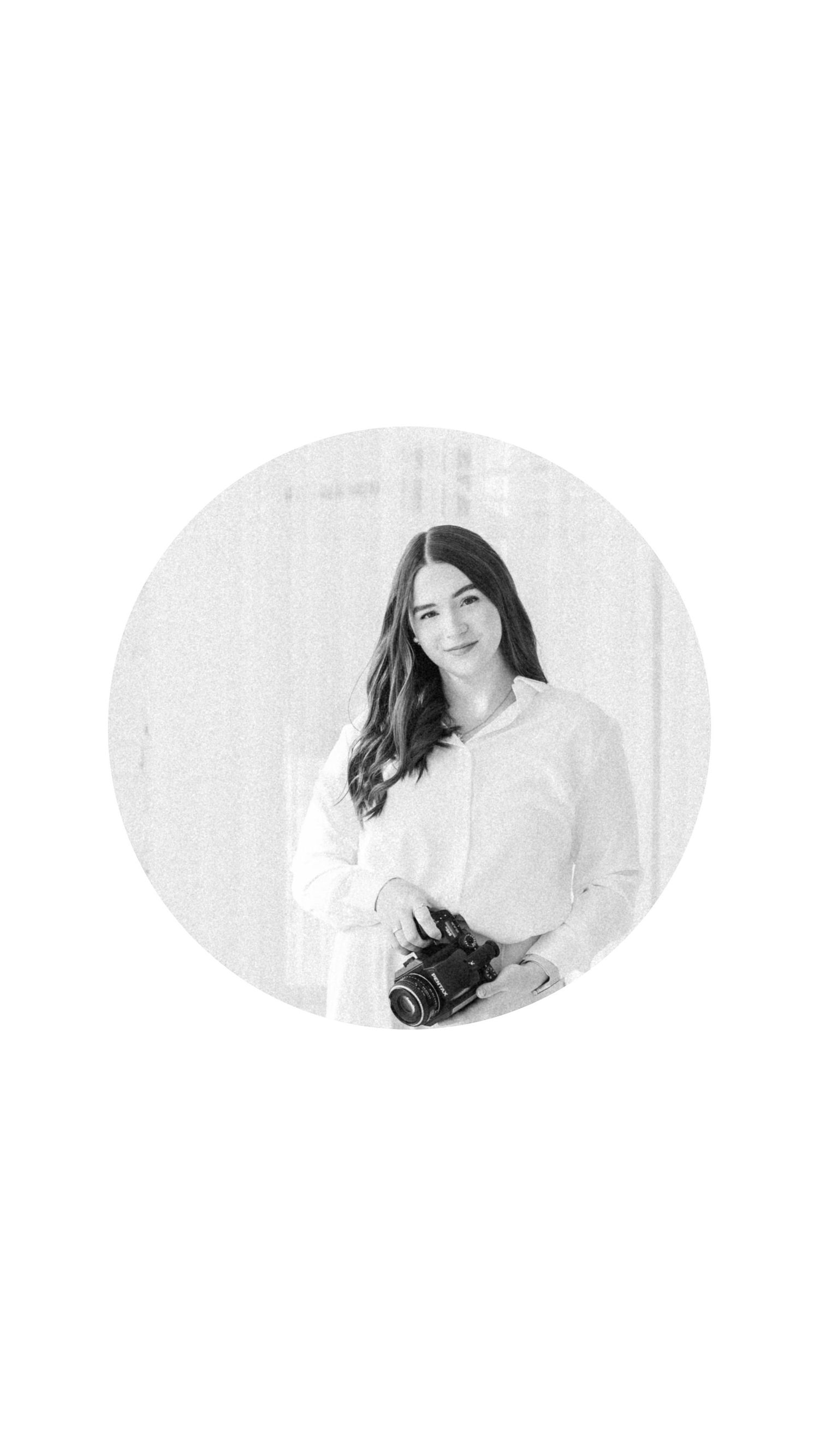Black and white portrait of a young woman holding a camera, wearing a white shirt, smiling softly.