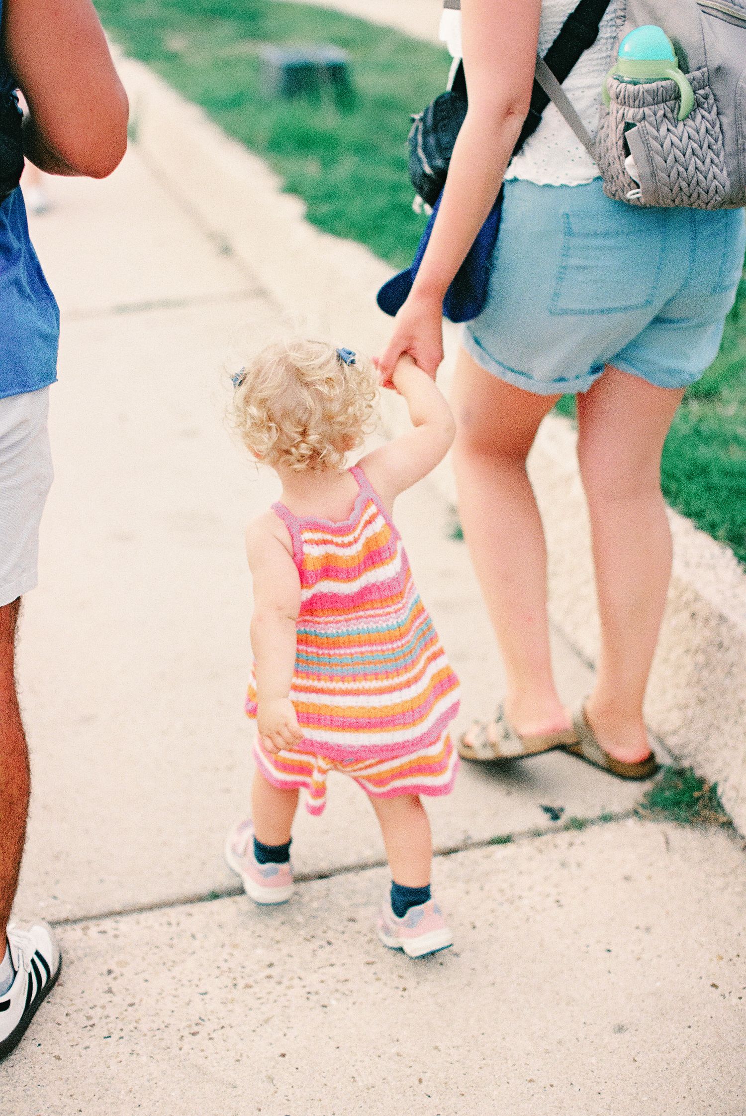 Toddler girl in colorful striped dress holds adult's hand while walking on sidewalk during sunny day.