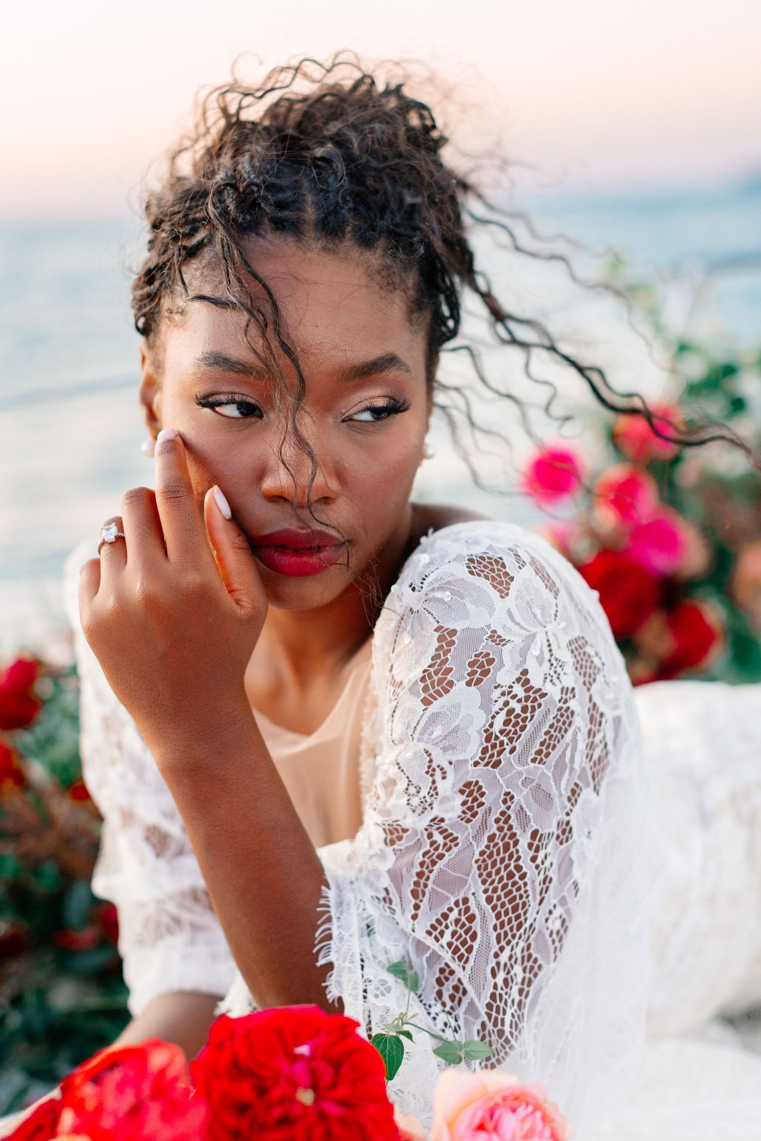 Beautiful Black bride in lace dress with red lips surrounded by roses near the sea.