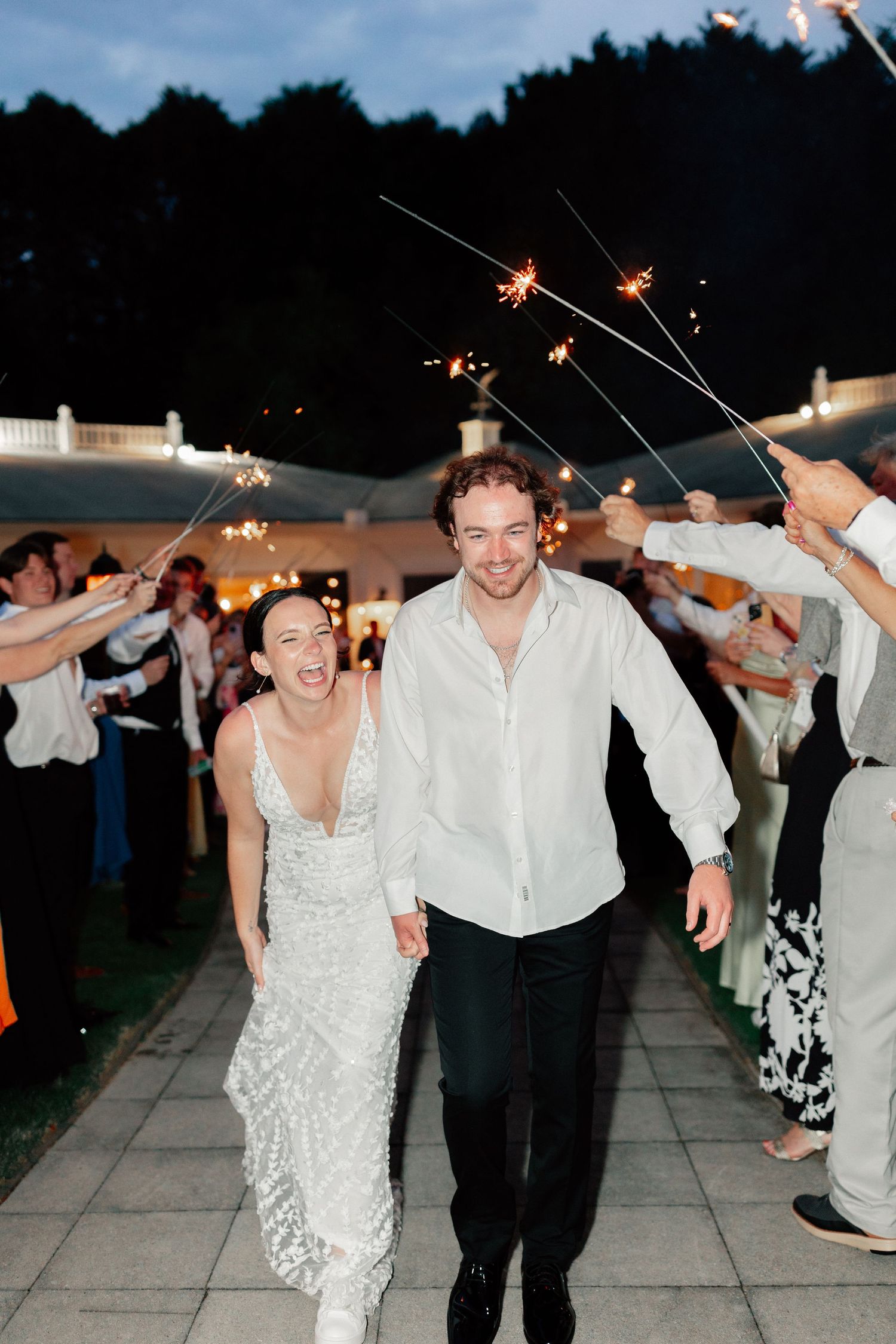 Bride and groom walk through sparkler tunnel at outdoor wedding reception at dusk, smiling joyfully.