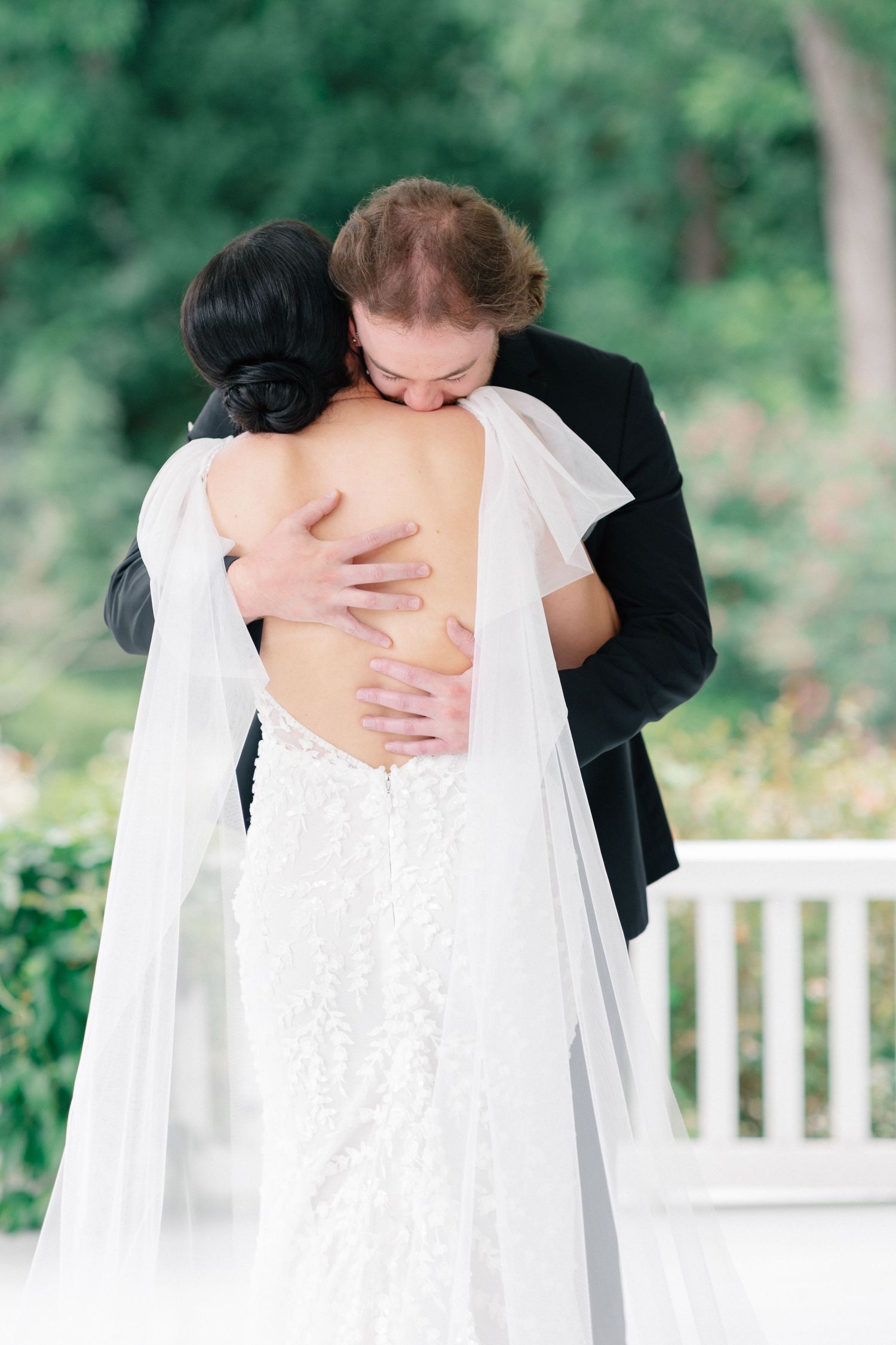 Bride and groom share an emotional first look hug, bride in backless lace gown with veil, outdoors.