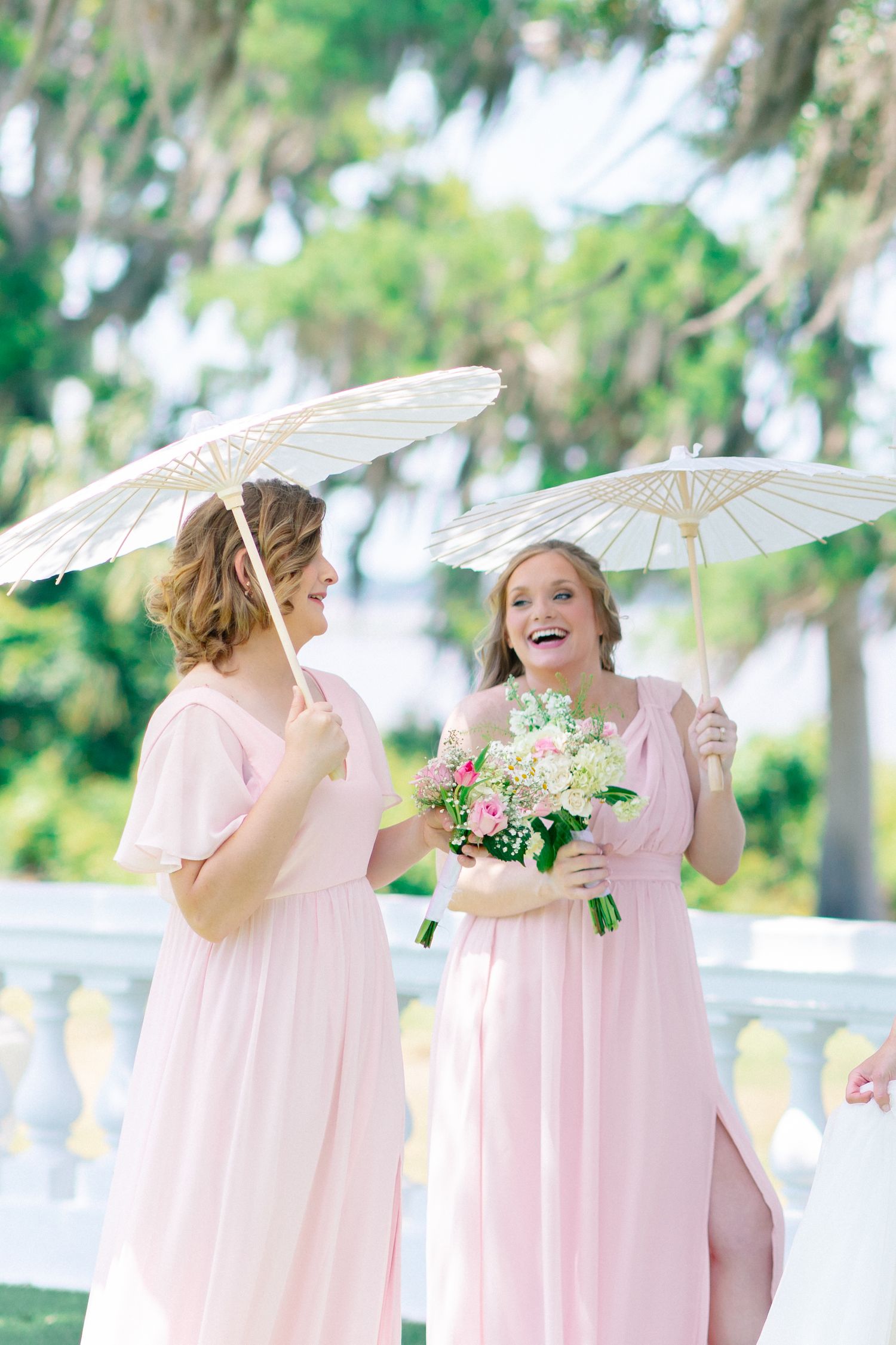 Two bridesmaids in pink dresses holding white parasols and a floral bouquet, laughing outdoors at a wedding, wedding at Bella Cosa