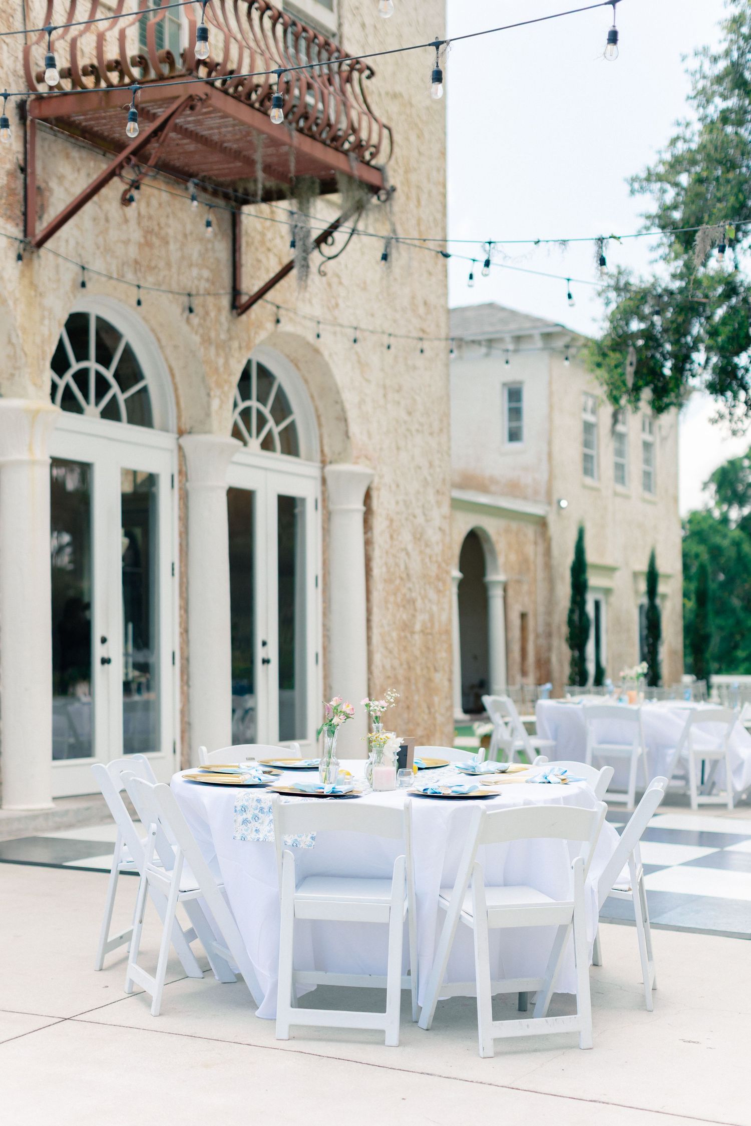 Elegant outdoor wedding reception with white round tables, folding chairs, string lights, and arched windows on a stone building, Bella Cosa