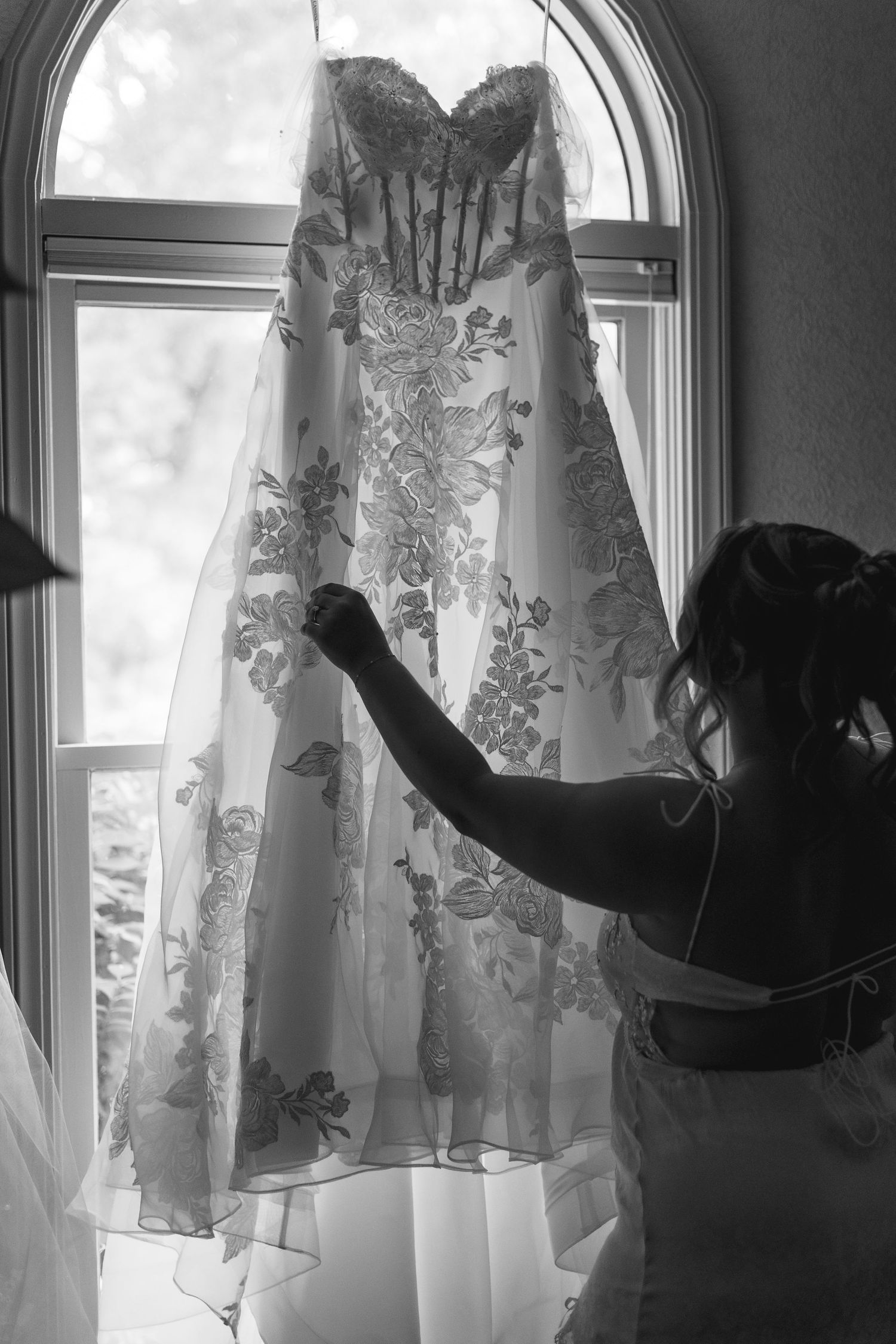 Black and white photo of a bride reaching up to touch her floral lace wedding gown hanging in a sunlit window.