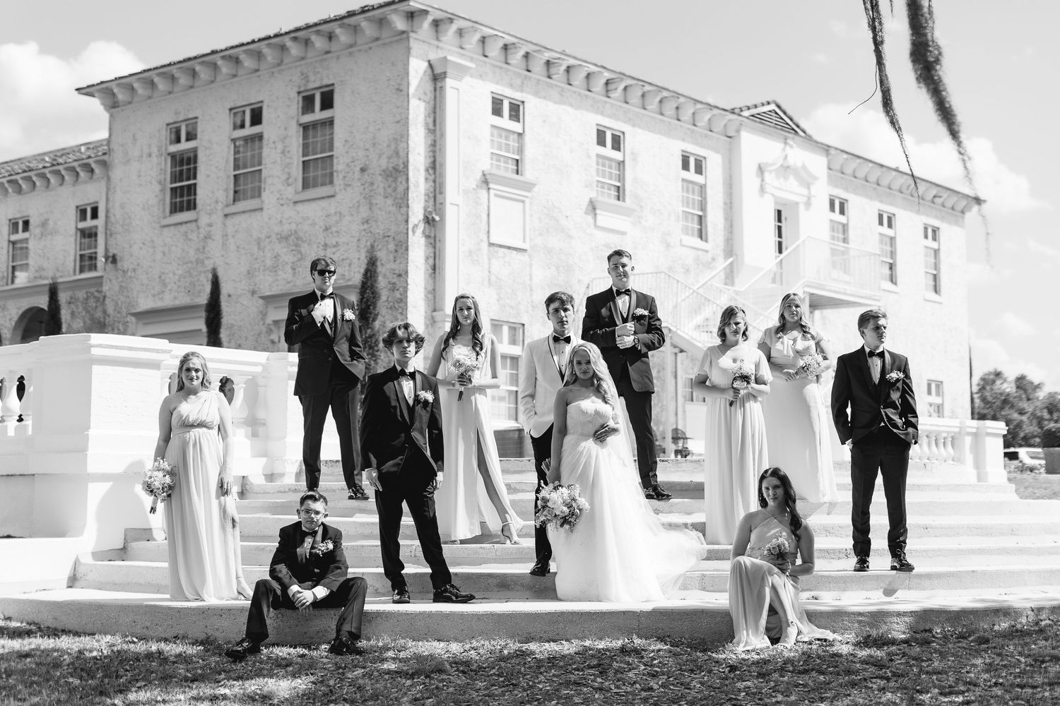 Black and white wedding party photo posed on grand estate steps with bride, groom, and elegantly dressed attendants.