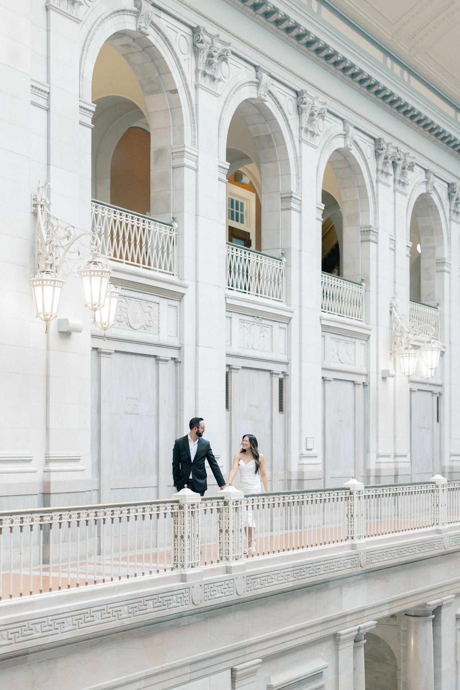 Couple holding hands walking along elegant white marble balcony with arched columns in grand neoclassical building.