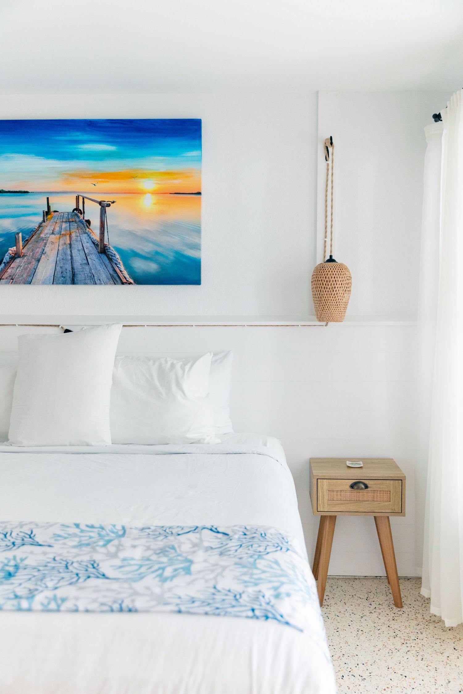 White beach bedroom with a wooden pier sunset canvas, rattan pendant light, and blue floral bedding on a white bed.