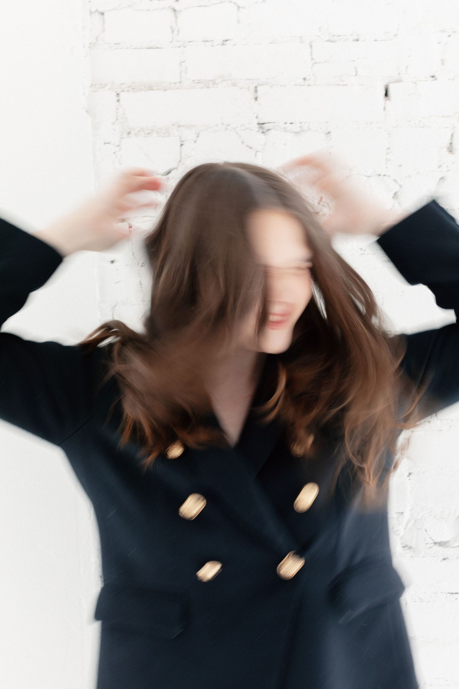 Woman in a black double-breasted blazer shakes her long brown hair in front of a white brick wall.
