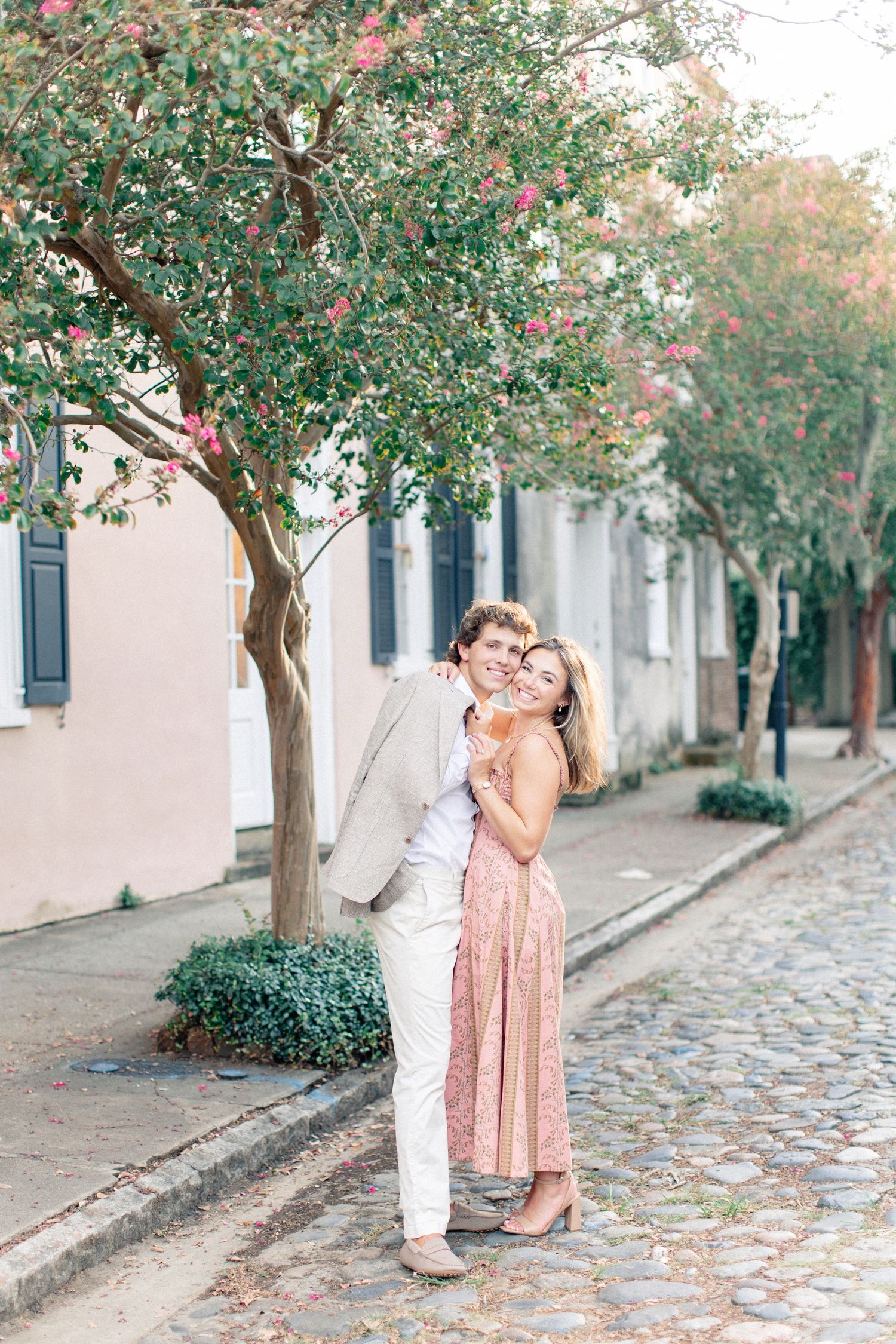 Couple embracing on a cobblestone street lined with blooming trees and colorful historic buildings.