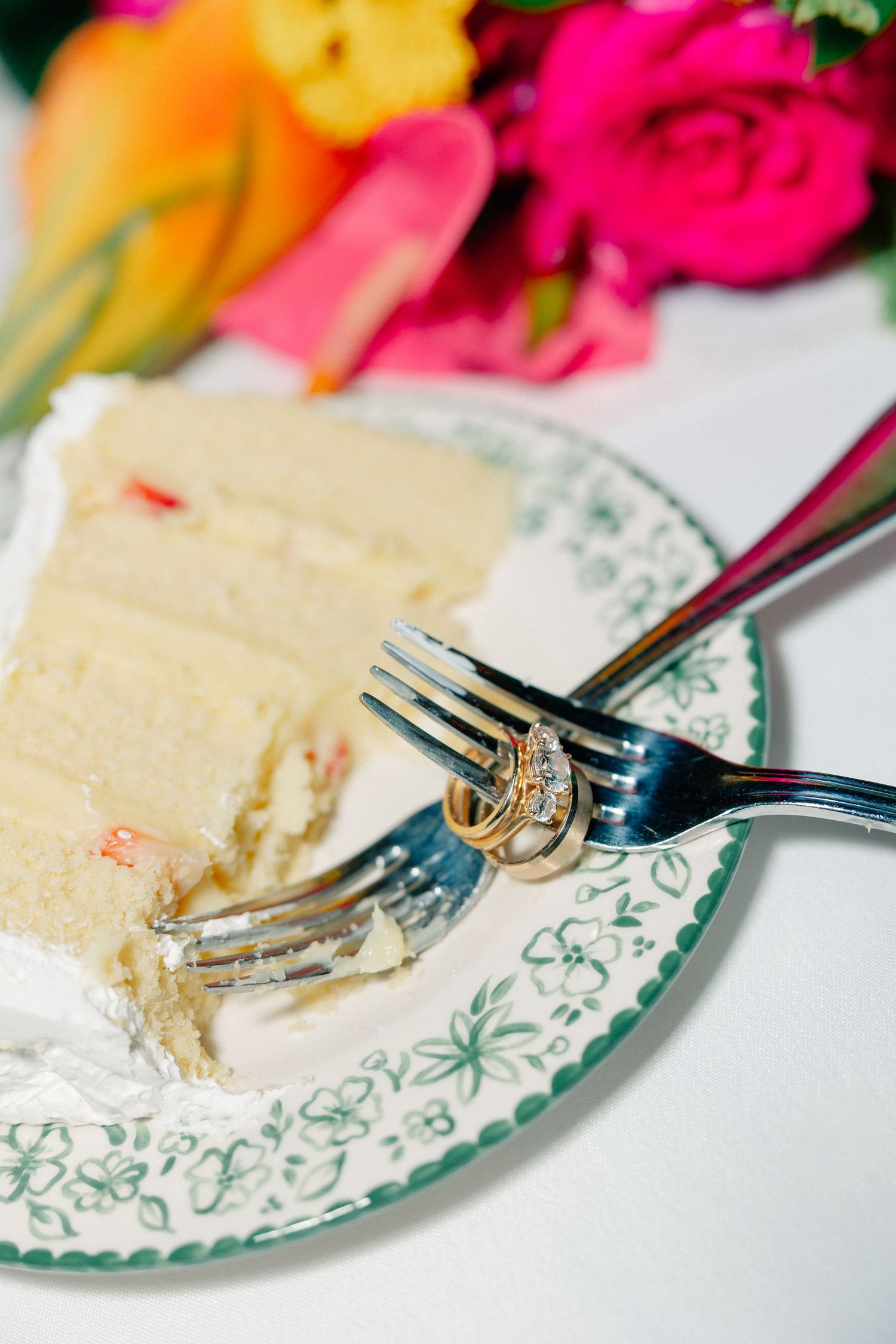 Wedding rings on a fork beside a slice of white layered cake on a floral plate with colorful flowers in background.