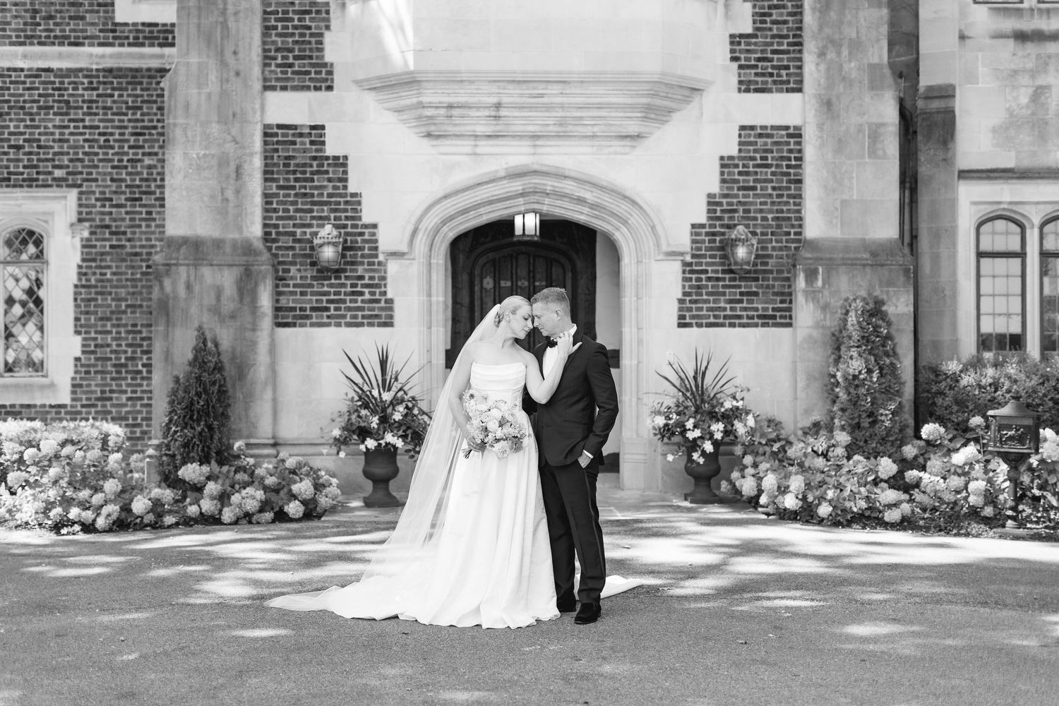 Bride and groom share a kiss in black and white portrait outside floral-adorned Tudor brick estate entrance.