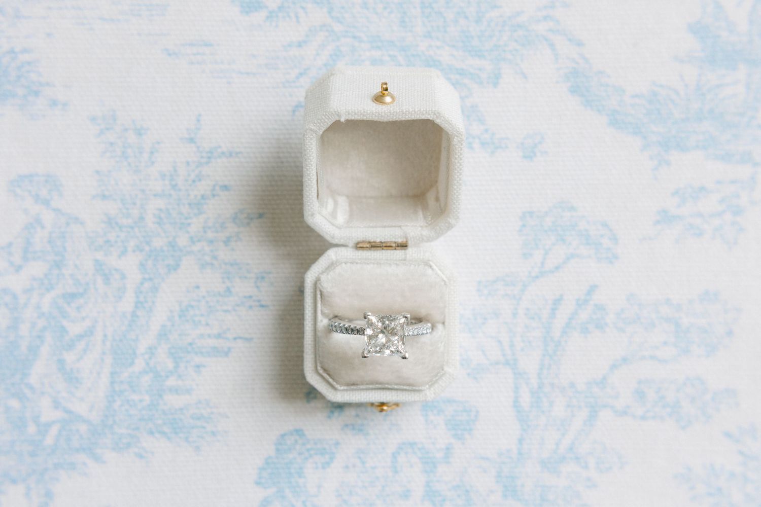 Diamond engagement ring in an open white velvet ring box on a blue toile background.