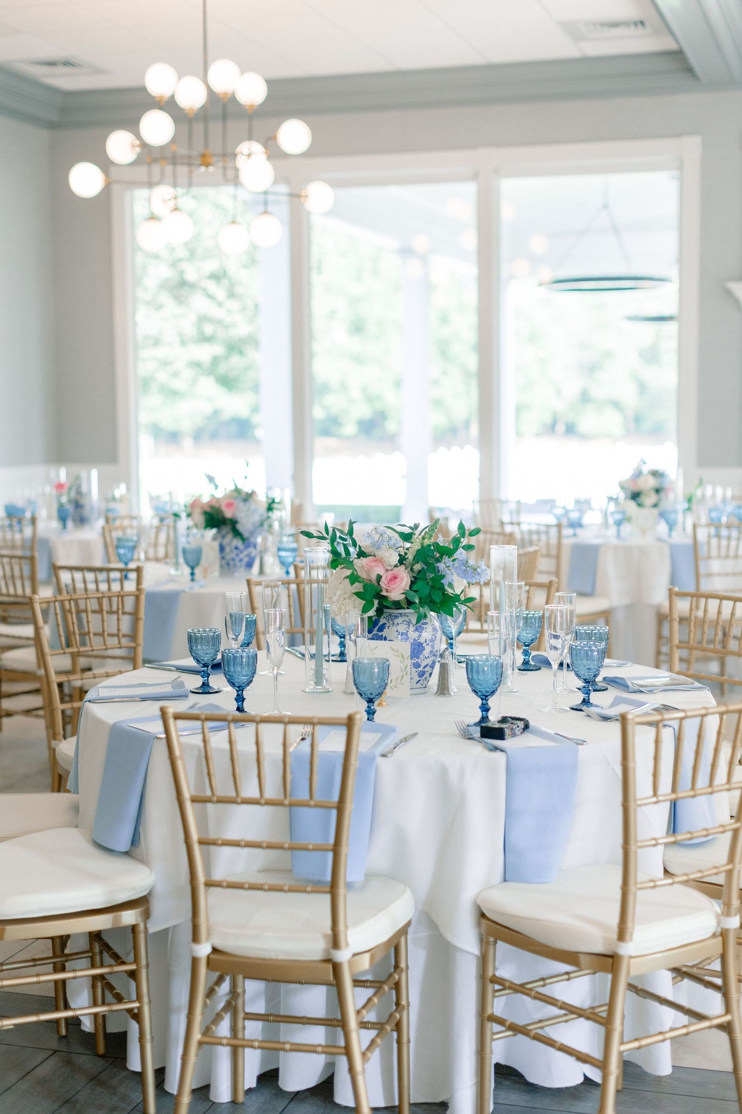 Elegant wedding reception with gold Chiavari chairs, blue napkins, floral centerpieces, and blue glassware on white linen tables.