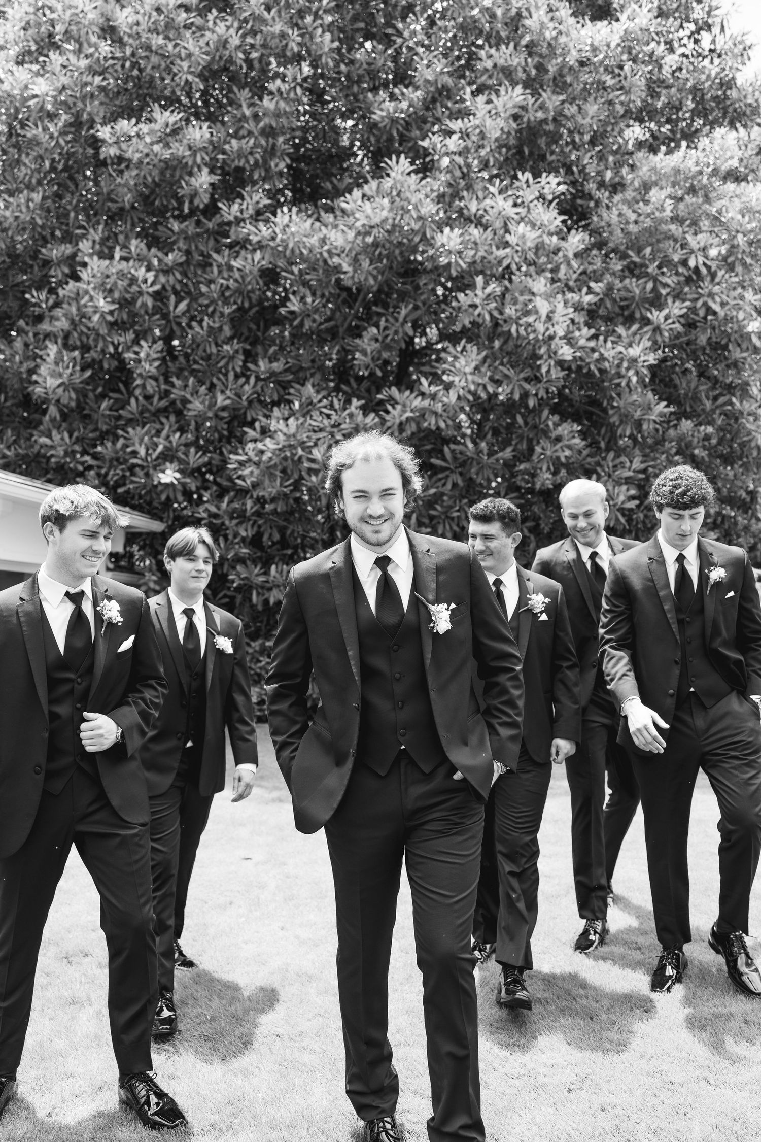 Groom leads groomsmen in dark suits with boutonnieres, walking outdoors in a classic black and white wedding photo.