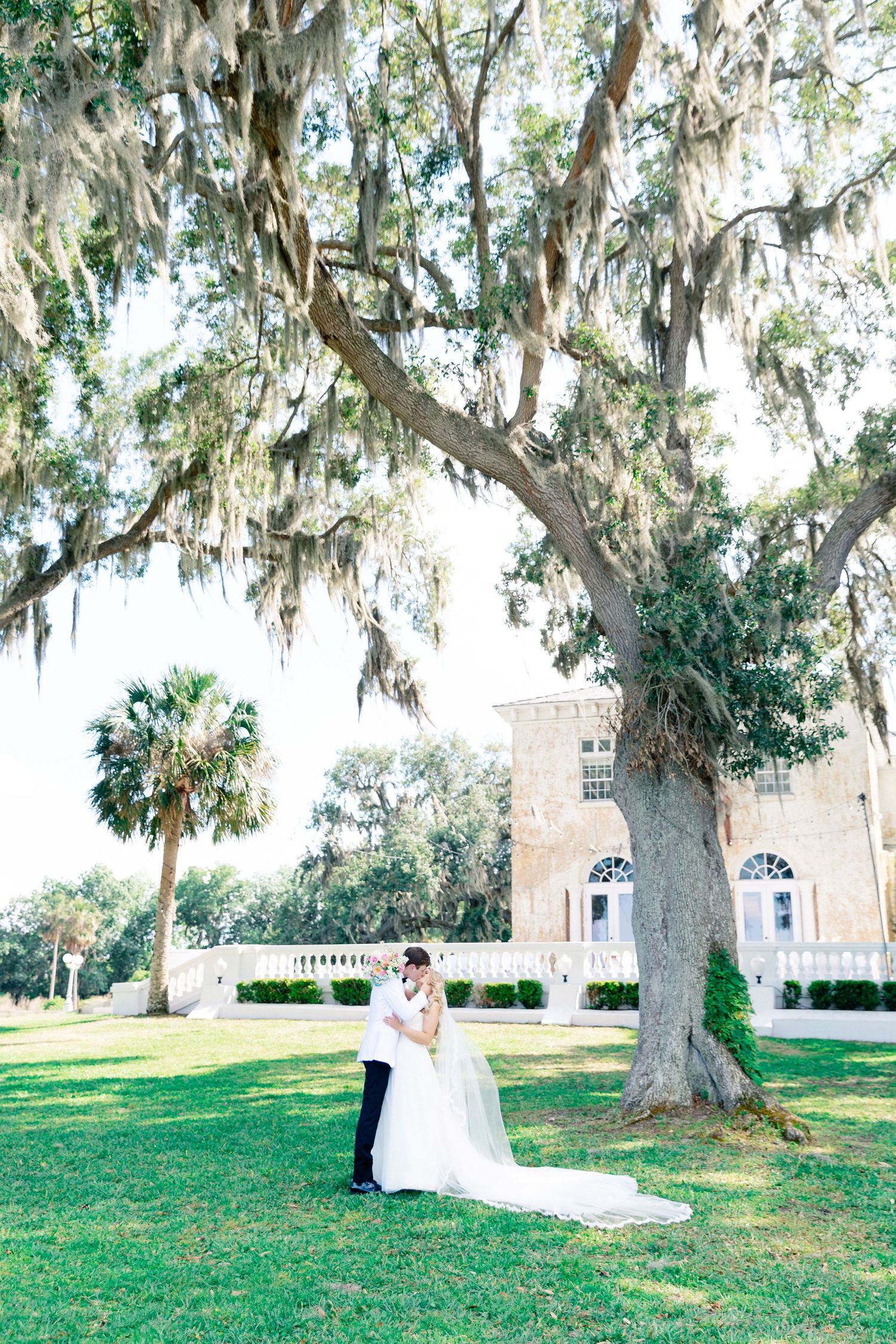 Bride and groom share a romantic moment under a moss-draped oak tree on lush green grounds of an elegant estate, wedding at Bella Cosa