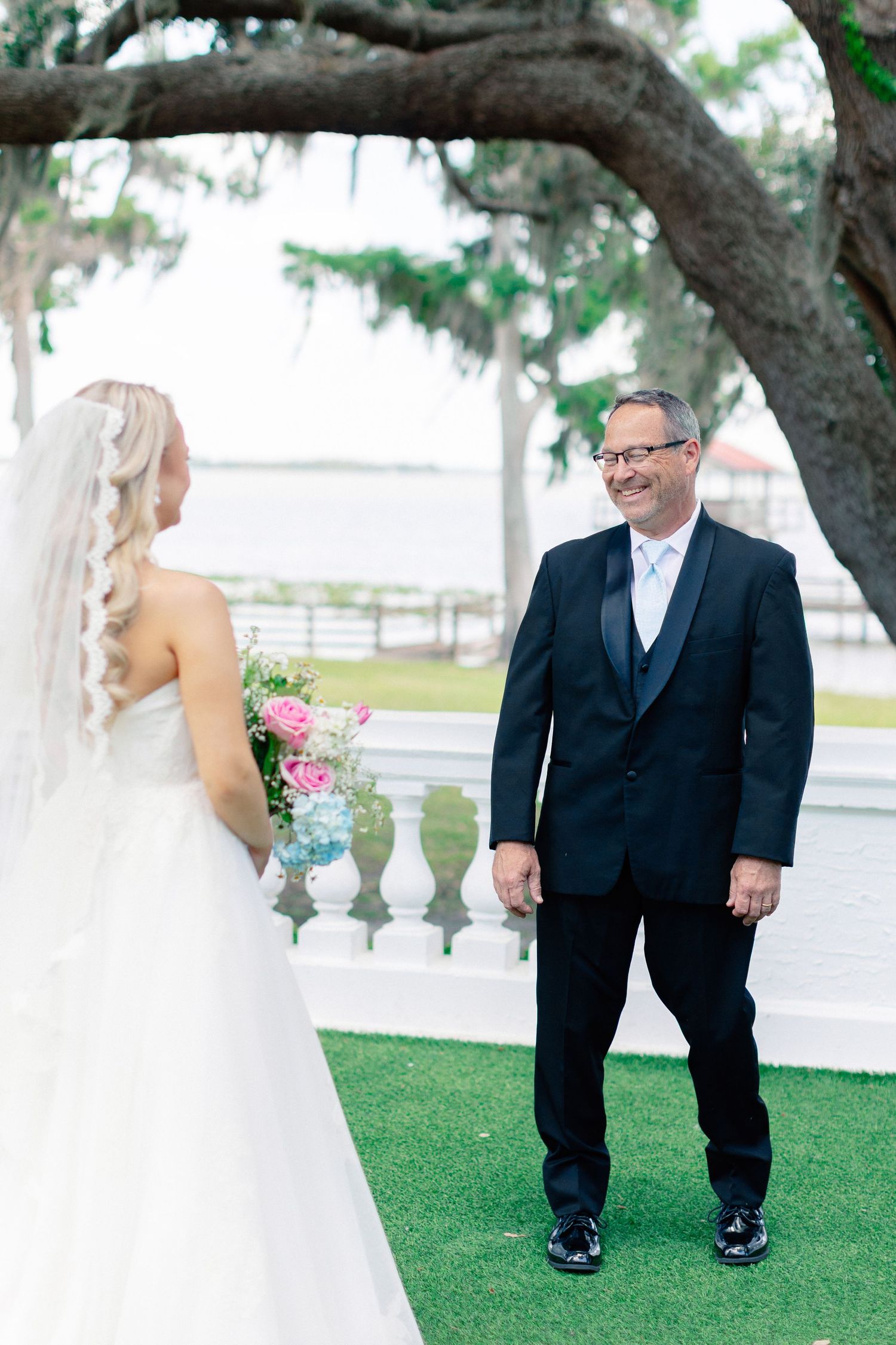 Groom in black tuxedo smiling at bride in white strapless gown holding pink bouquet during outdoor first look, wedding at Bella Cosa
