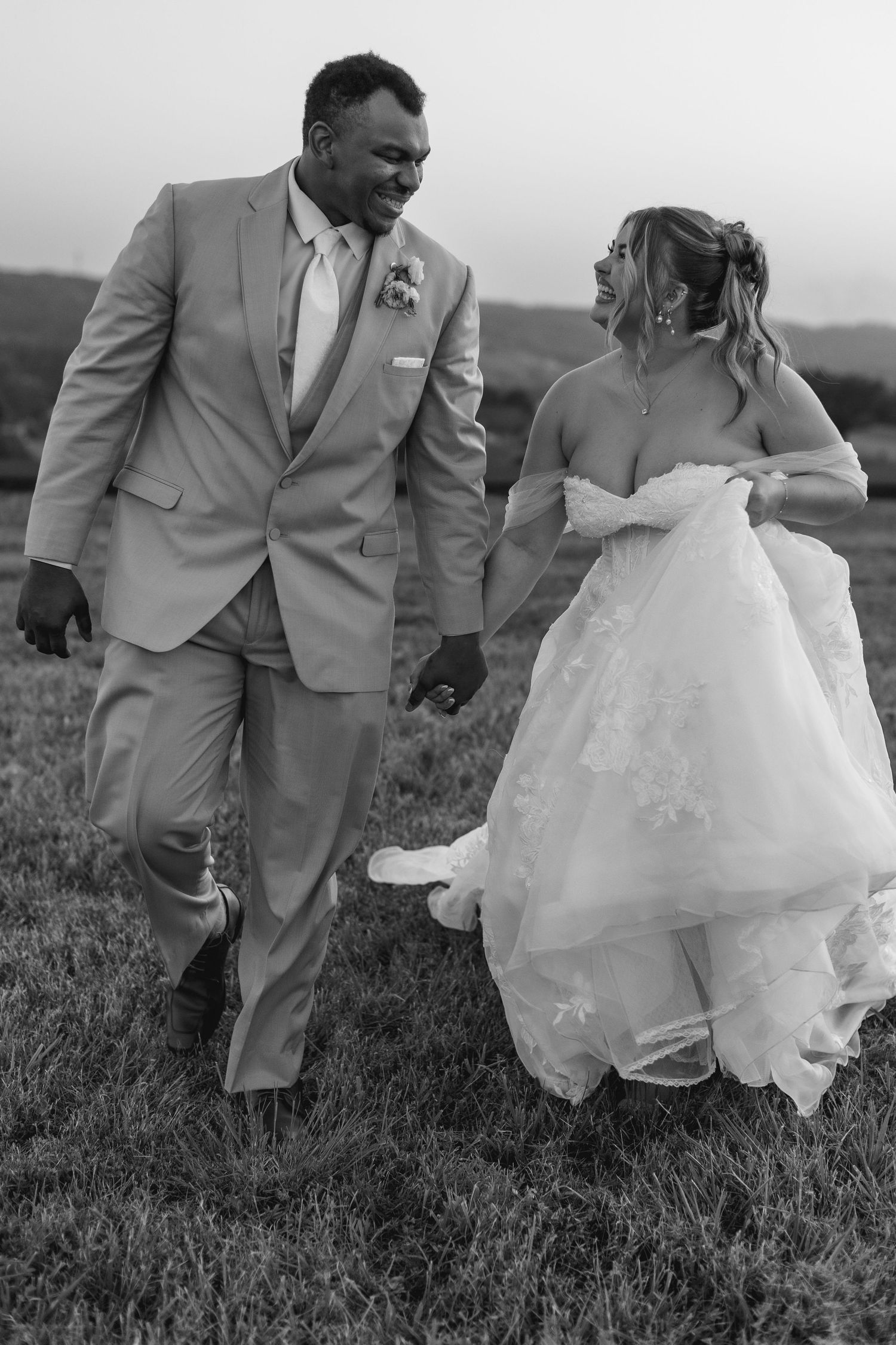Black and white photo of smiling newlyweds strolling through field, groom in tan suit, bride lifting white gown.