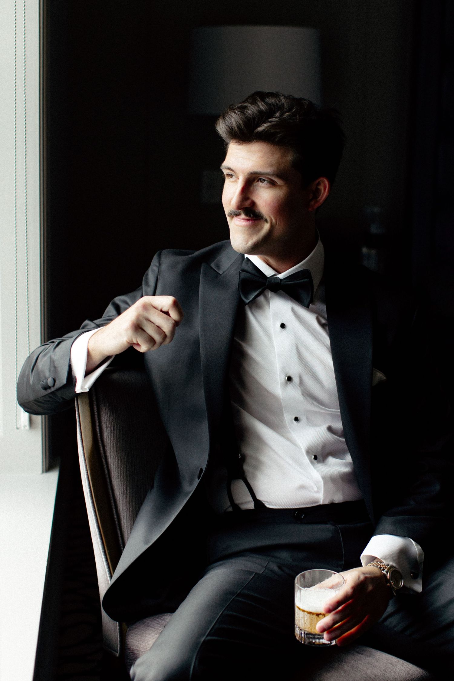 Elegant man in black tuxedo with bow tie sitting in chair holding whiskey glass, smiling confidently near window.