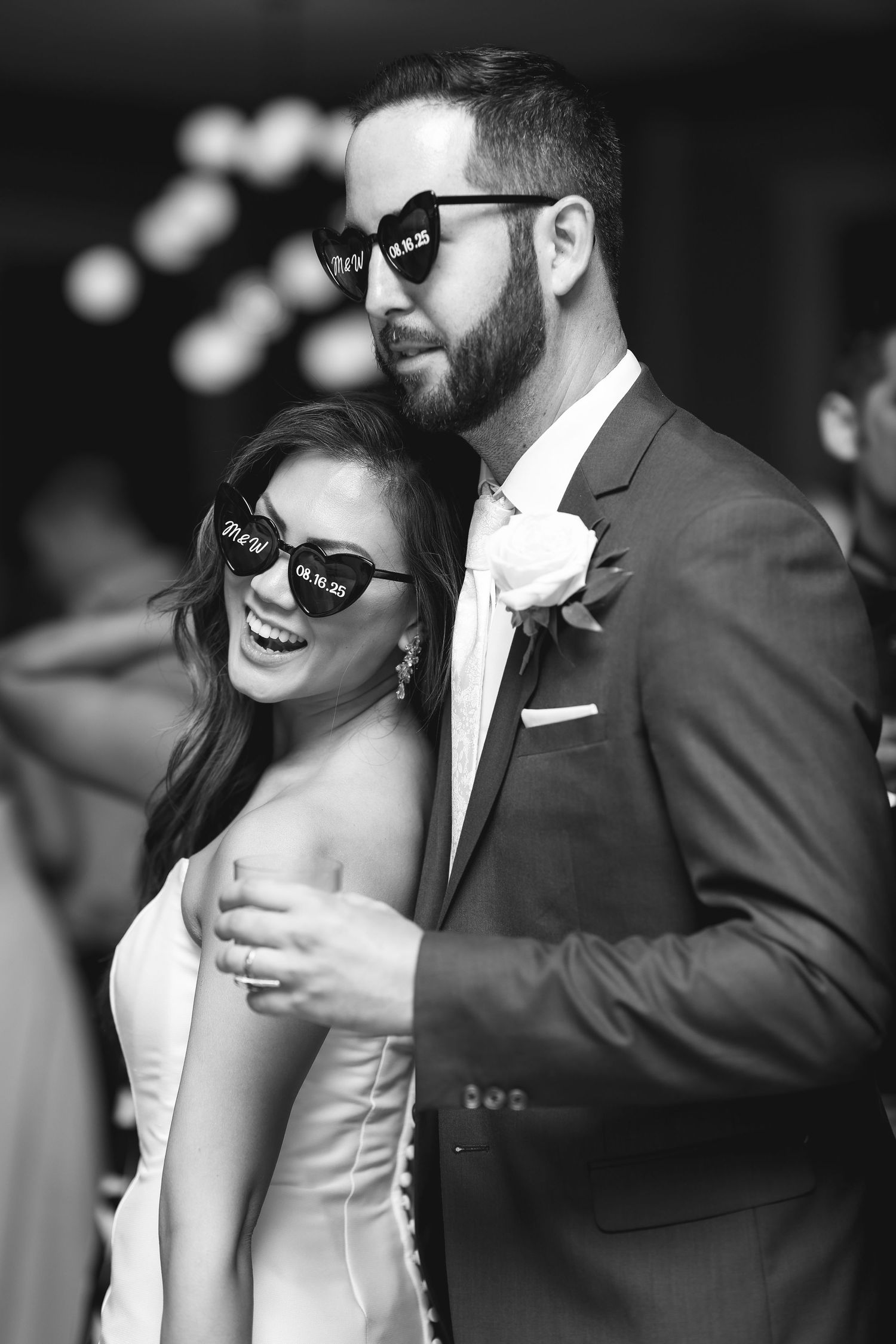 Black and white photo of smiling bride and groom wearing novelty sunglasses dancing together at their wedding reception.