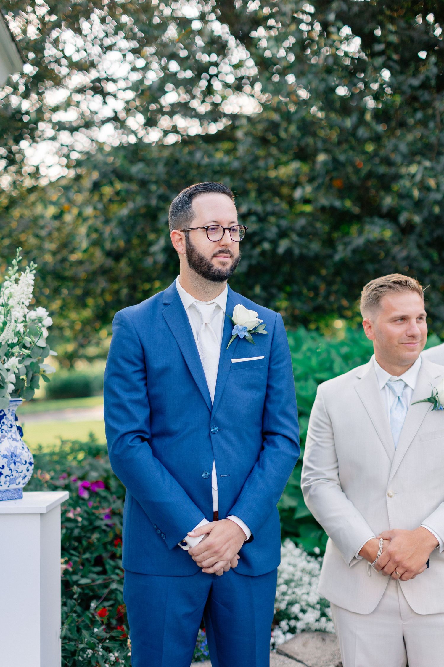 Groom in blue suit with white boutonniere stands at outdoor wedding altar beside groomsman in beige suit.