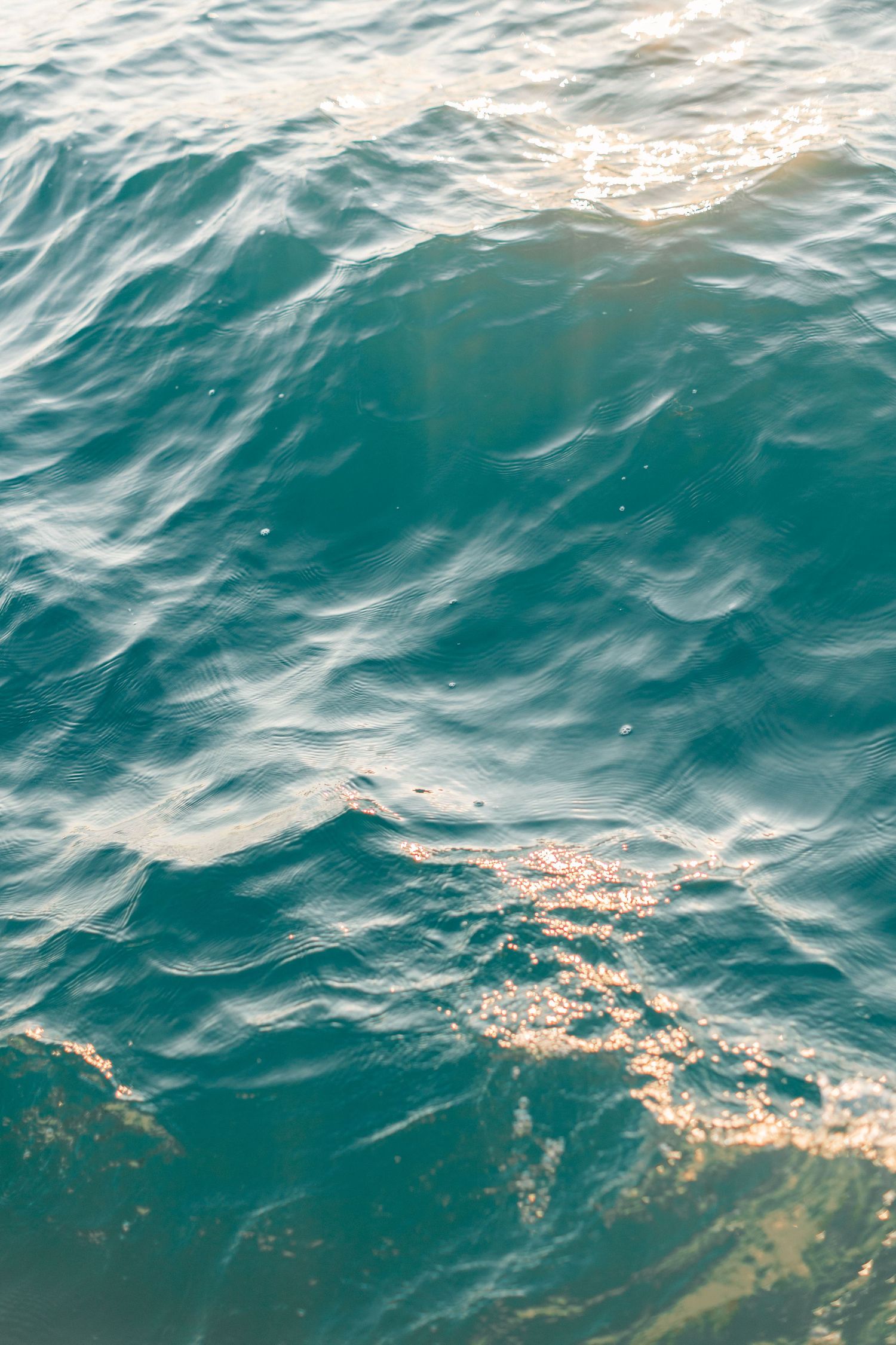 Sunlight glistening on rippling teal ocean water surface with golden reflections creating a serene sea texture background.