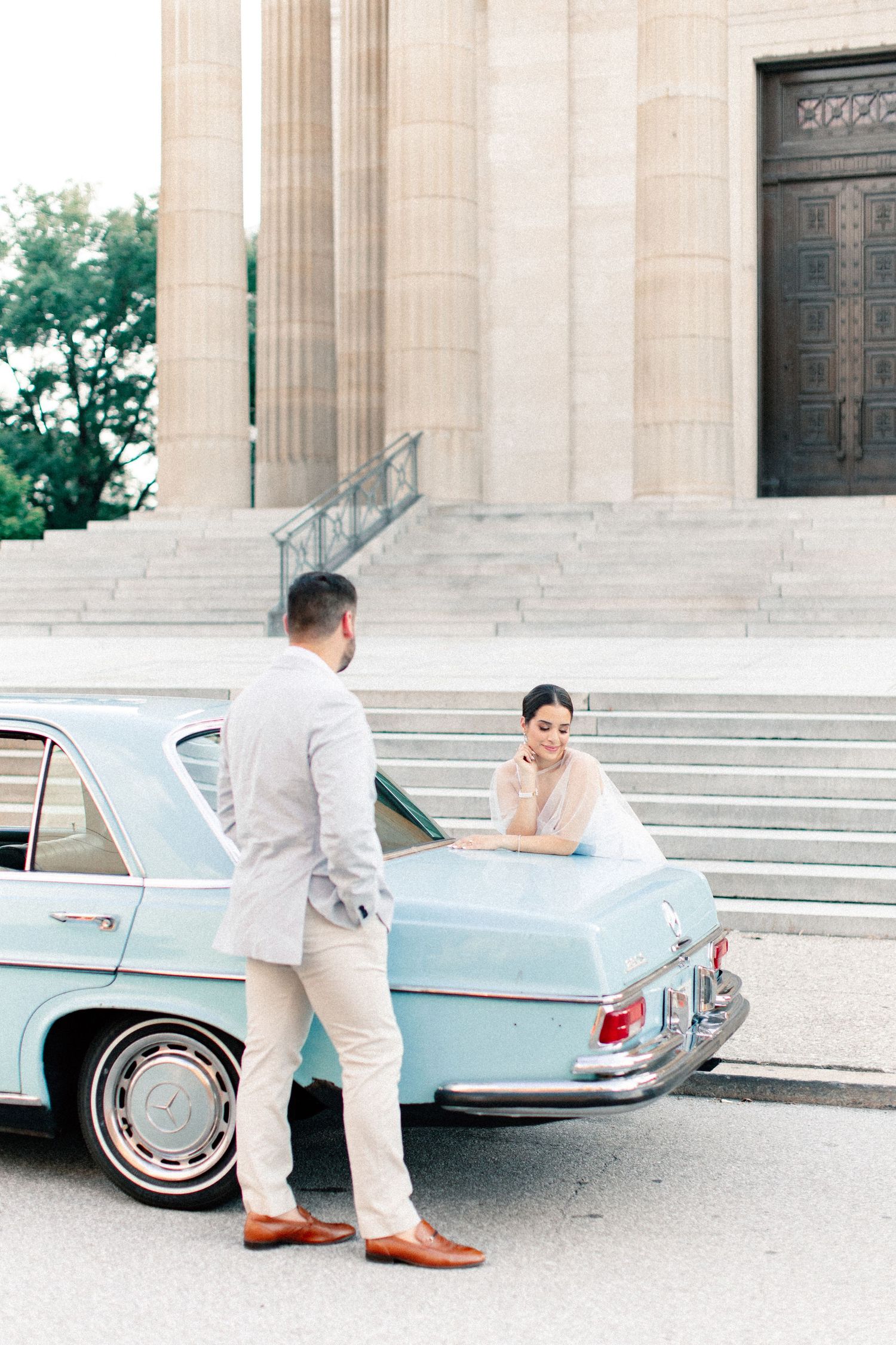 Bride in white dress leans on vintage light blue Mercedes at neoclassical building steps with groom in gray suit.