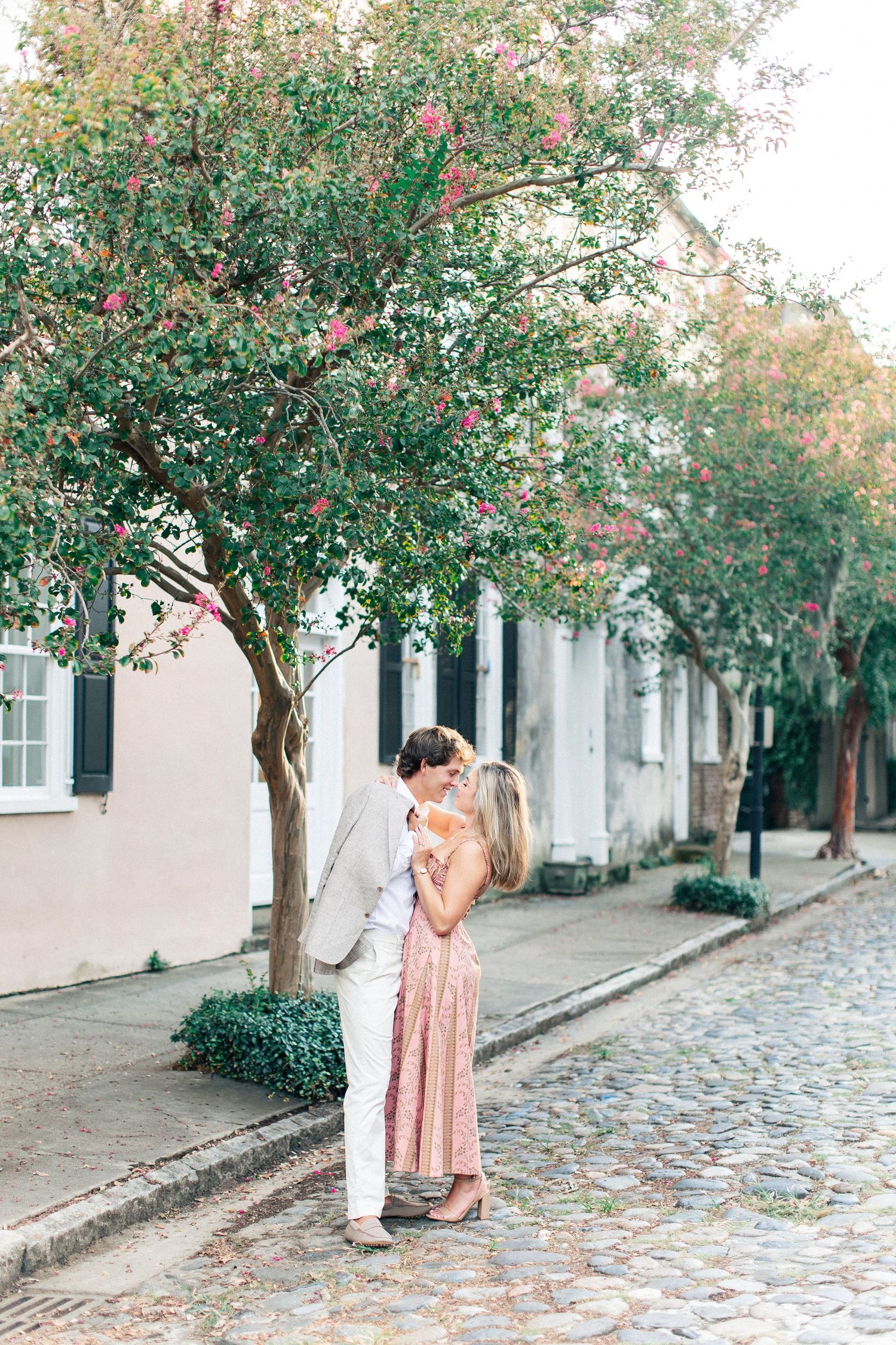 A couple shares a tender kiss on a cobblestone street lined with blooming crepe myrtle trees.