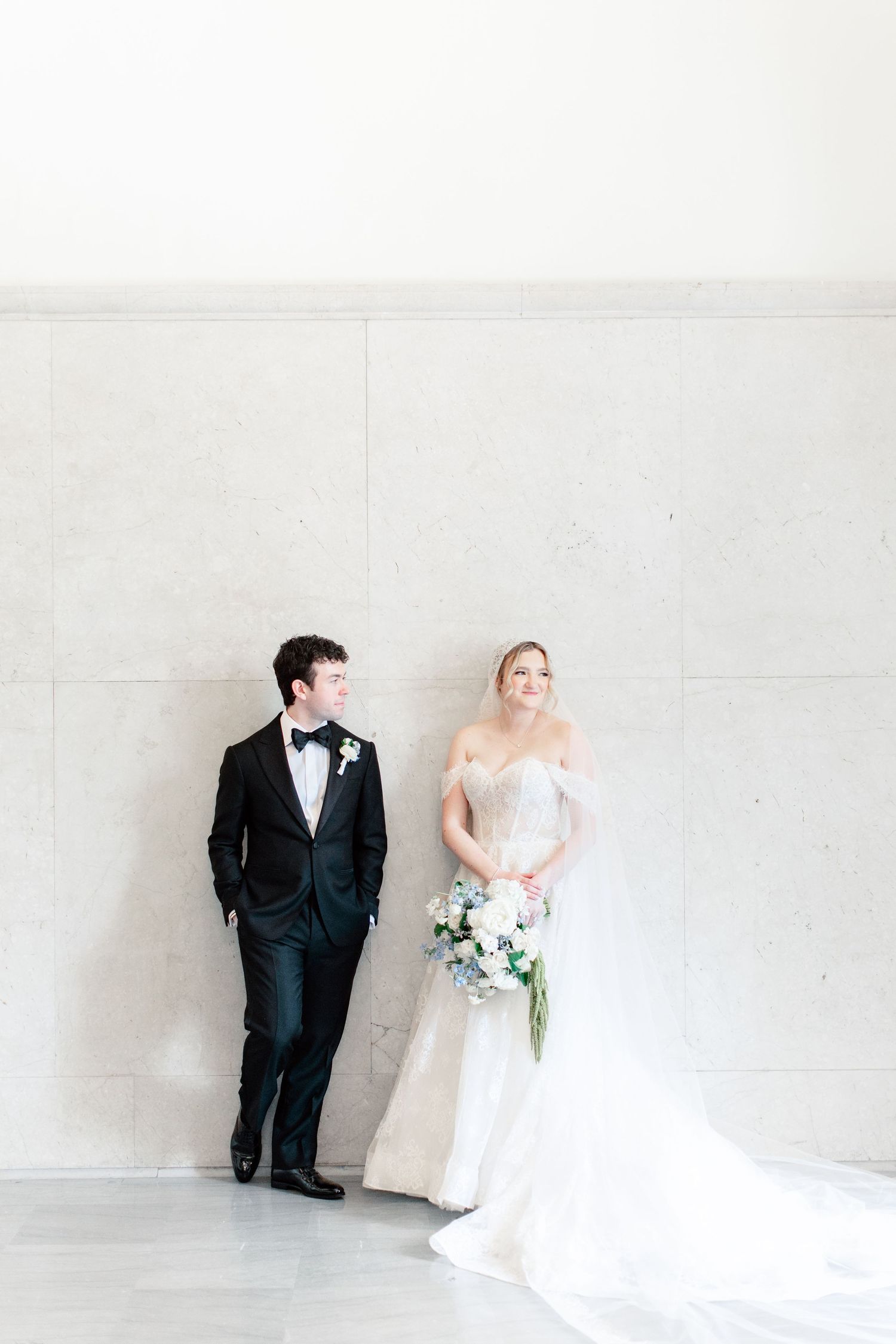 Bride and groom lean against a marble wall, she holds a blue and white bouquet in an off-shoulder gown, he wears a black tuxedo.