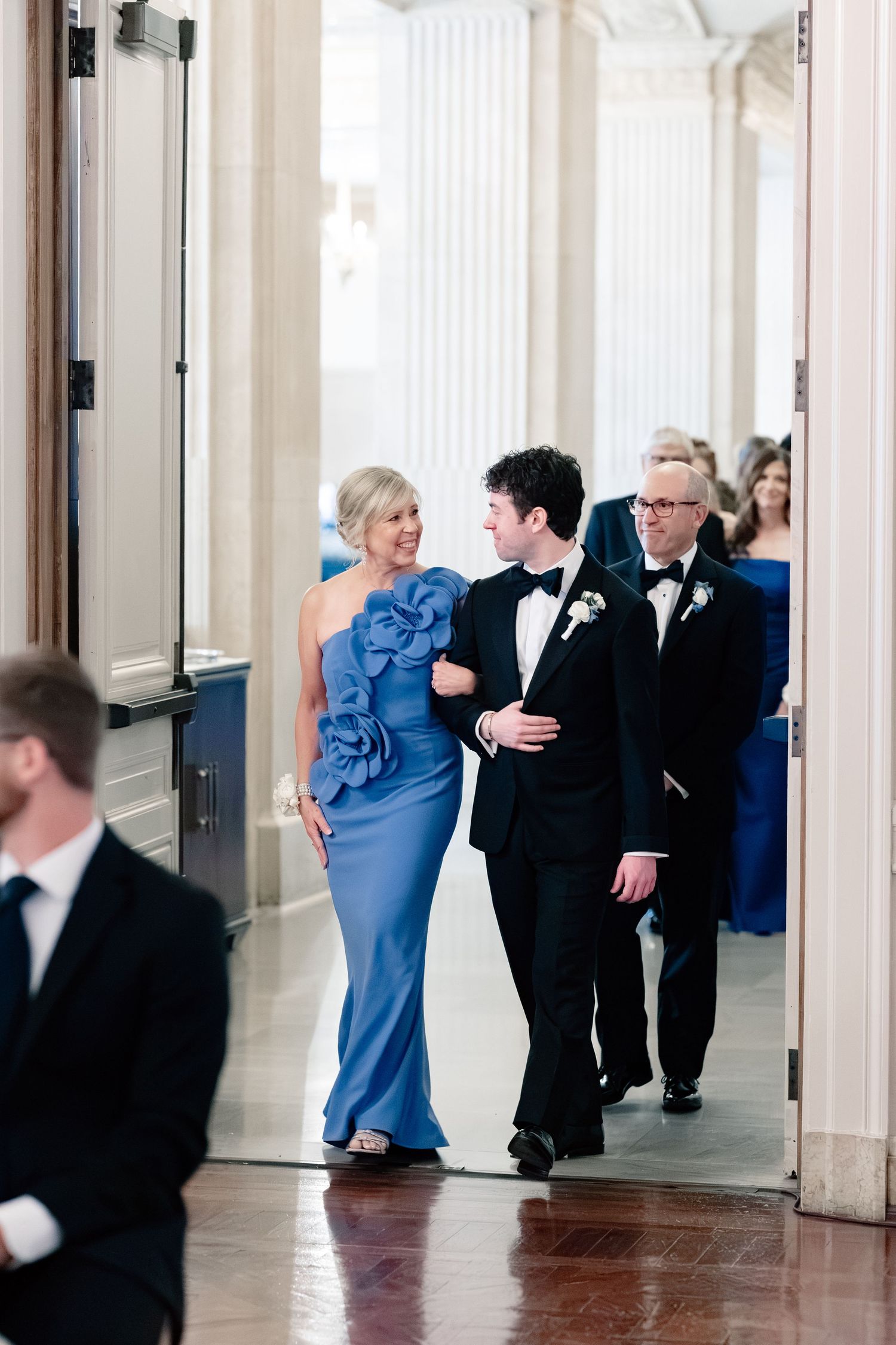 Groom in black tuxedo walks with elegantly dressed woman in blue gown through grand venue hallway.