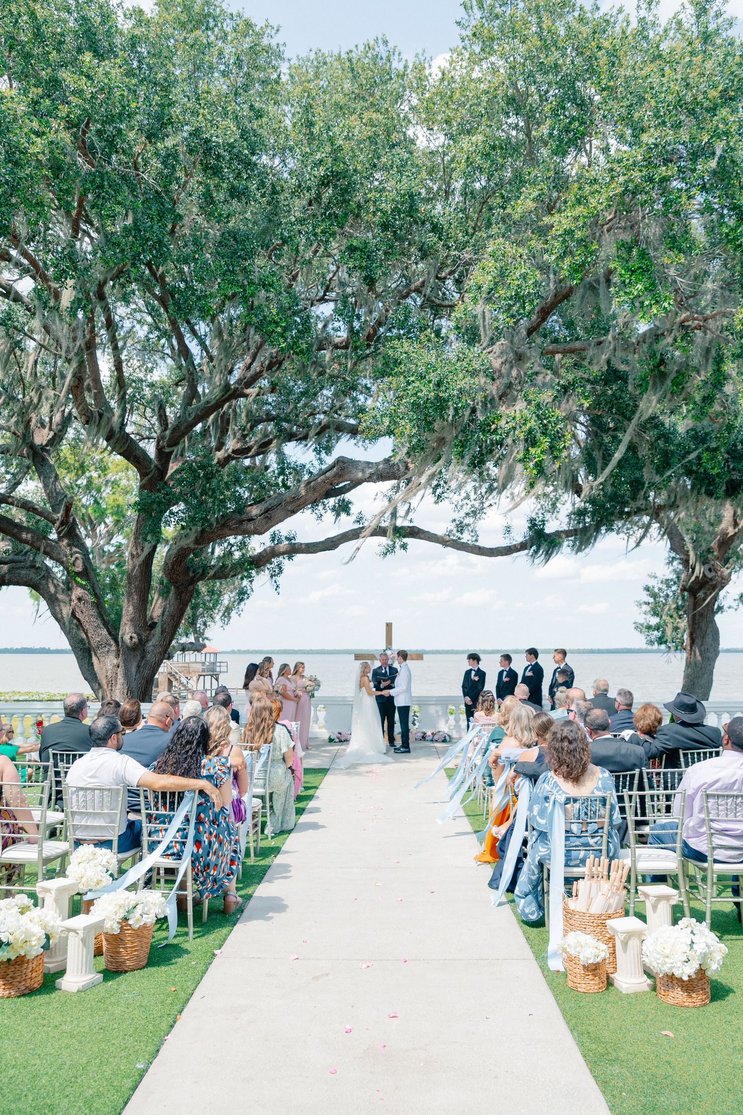 Outdoor waterfront wedding ceremony under large oak trees with guests seated along a white aisle runner, wedding at Bella Cosa