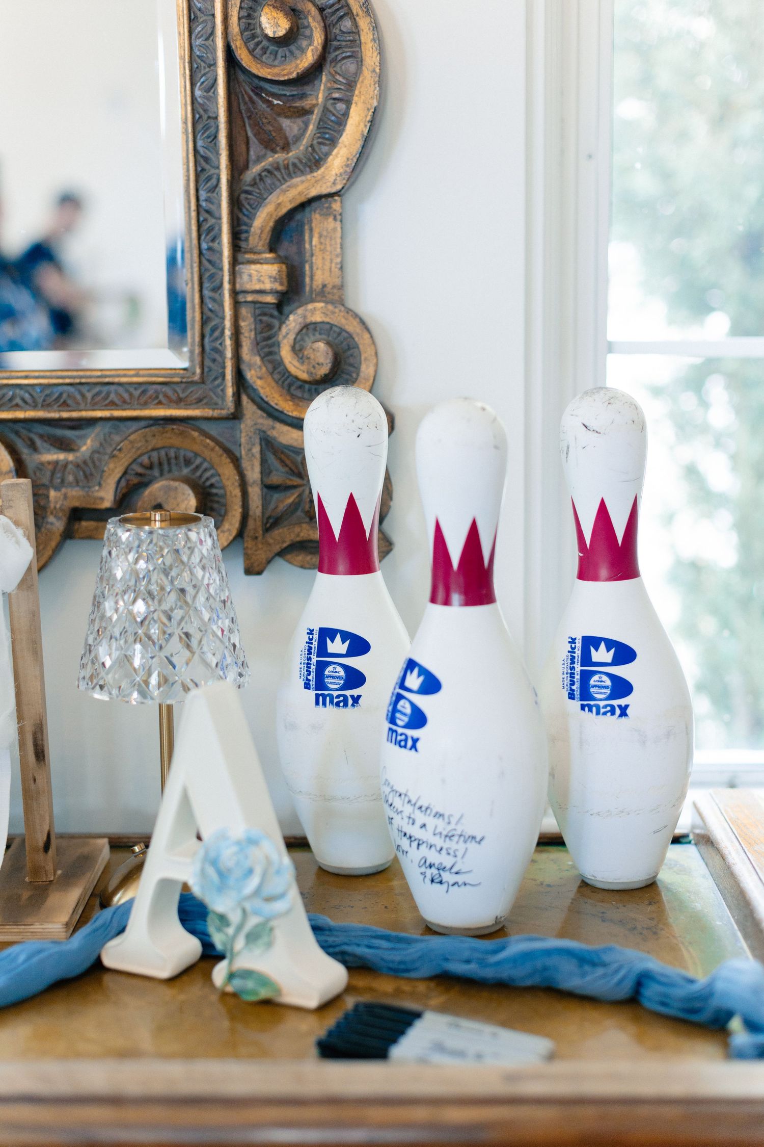 Three white B Max bowling pins with red crowns displayed on a wooden table beside a crystal lamp and ornate gold mirror, Bella Cosa
