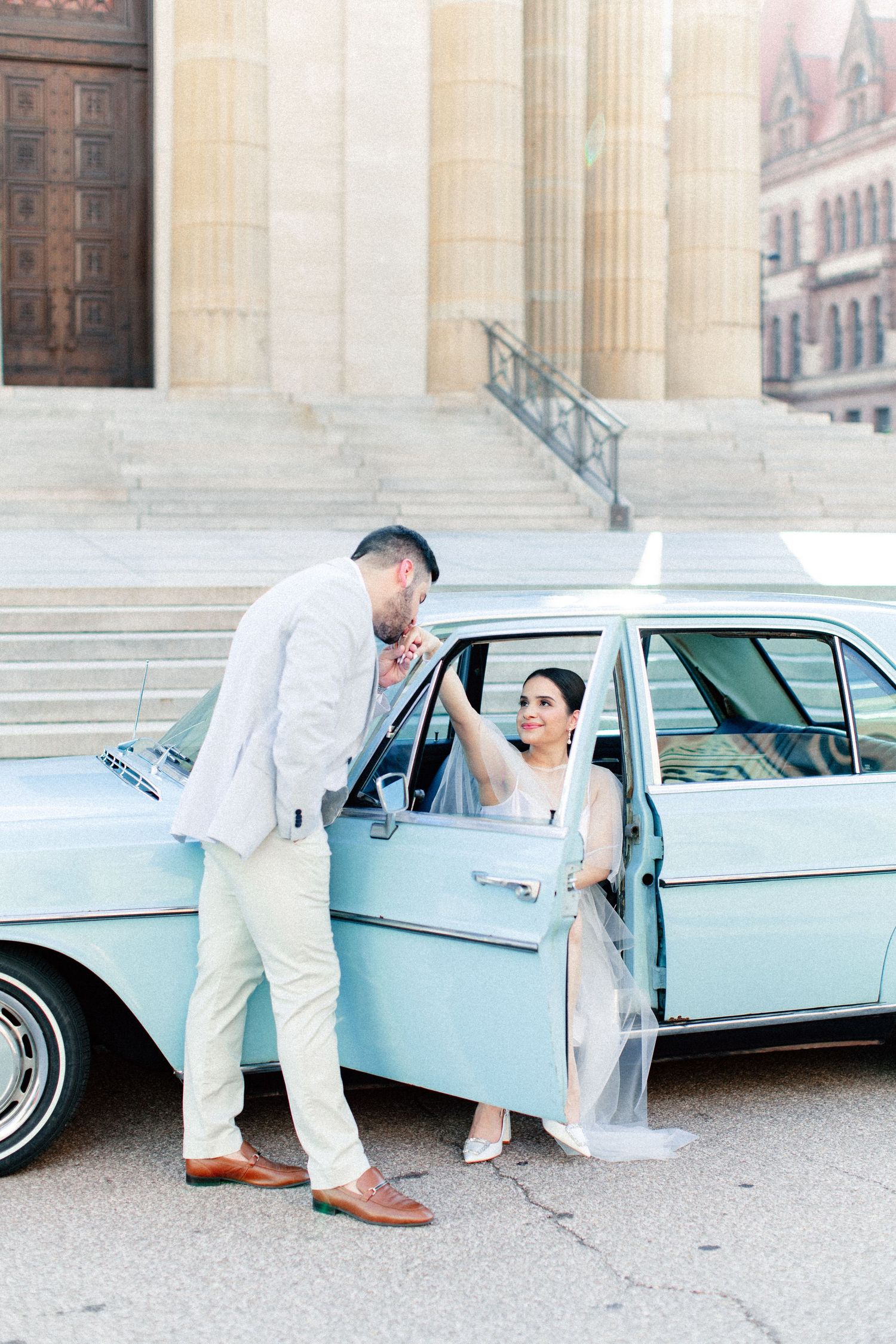 Bride and groom pose beside a vintage light blue car in front of a grand building on their wedding day.