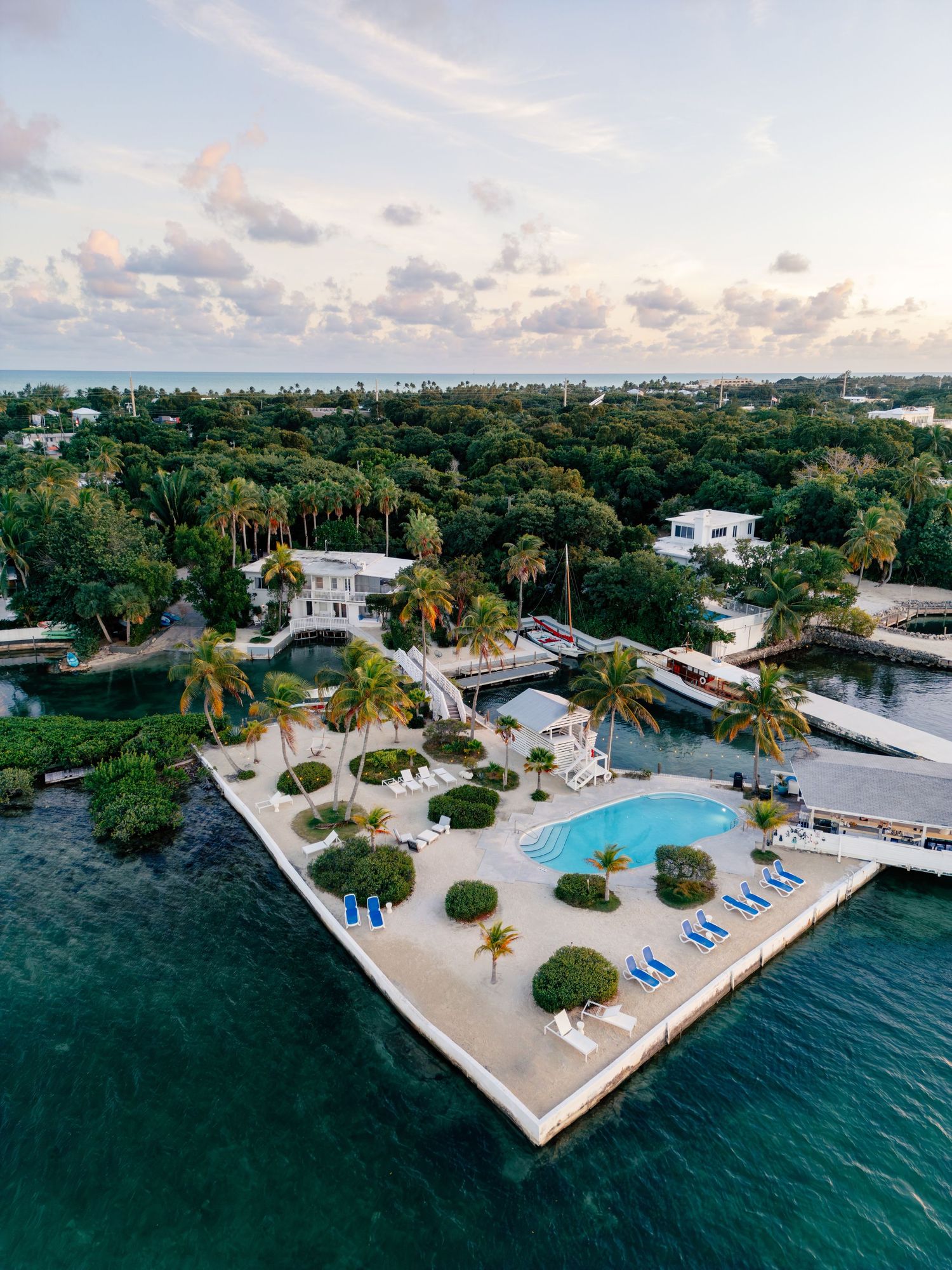 Aerial view of a luxury island resort with a pool, palm trees, and blue lounge chairs surrounded by turquoise water.