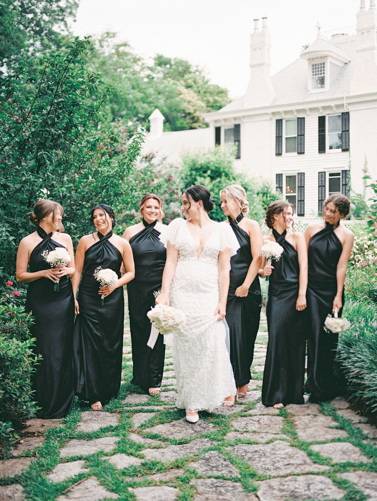 Bride in white lace gown stands with six bridesmaids in black halter dresses holding white bouquets outside elegant estate.