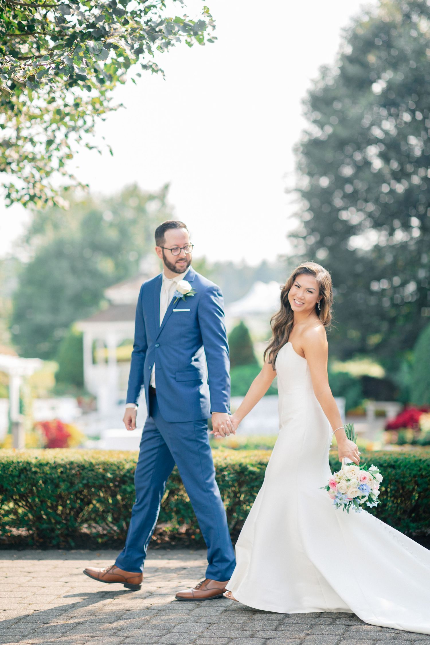 Bride in white gown and groom in blue suit walk hand in hand through a garden on their wedding day.