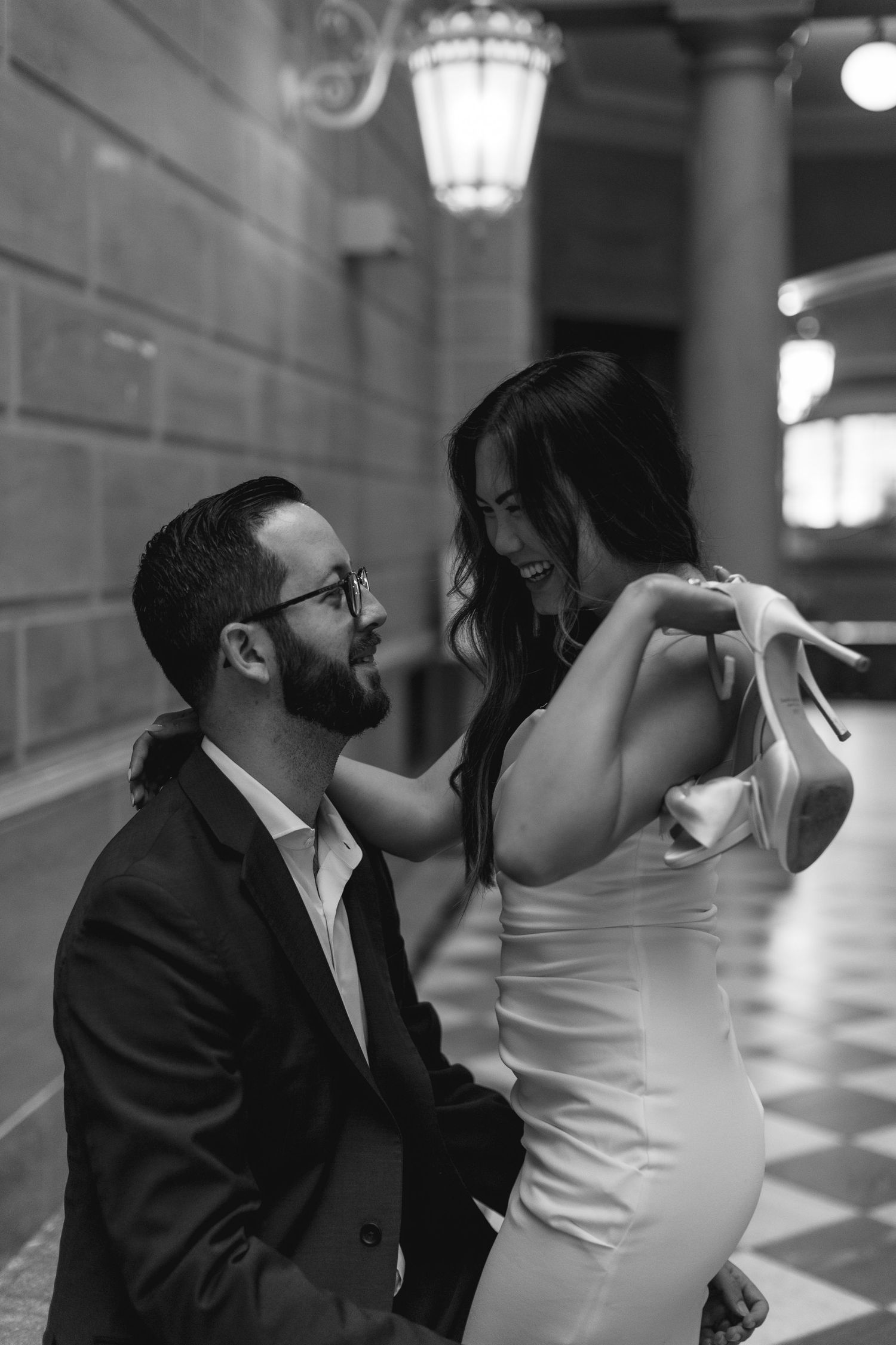Black and white photo of a groom kneeling before his smiling bride holding heels in an elegant columned hall.