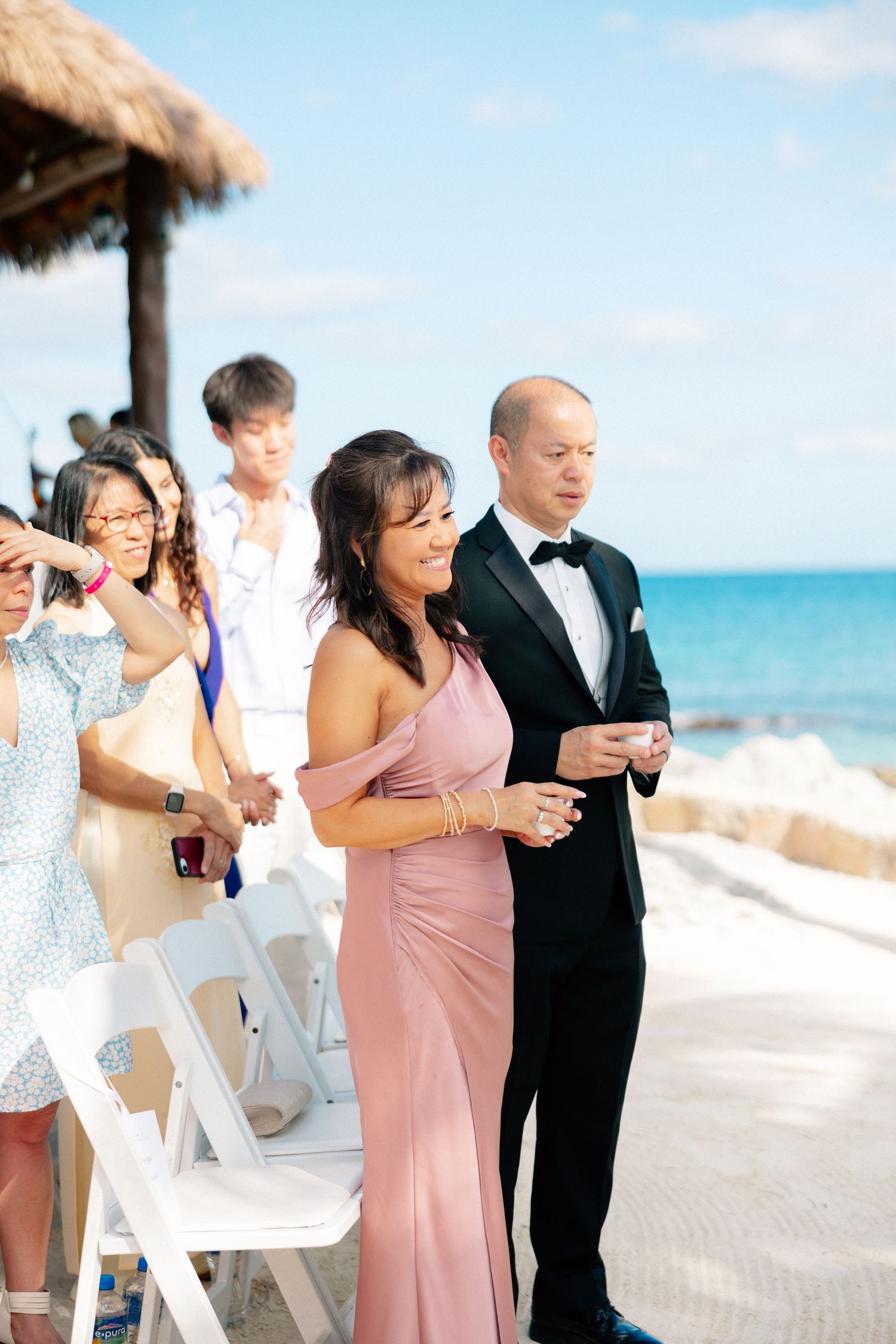 Couple in formal attire walk down beach wedding aisle, woman in pink gown, man in tuxedo, ocean backdrop.
