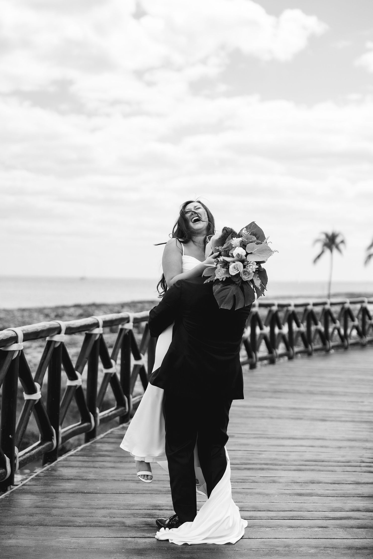 Groom lifting laughing bride on seaside boardwalk, black and white wedding photo with palm tree backdrop.