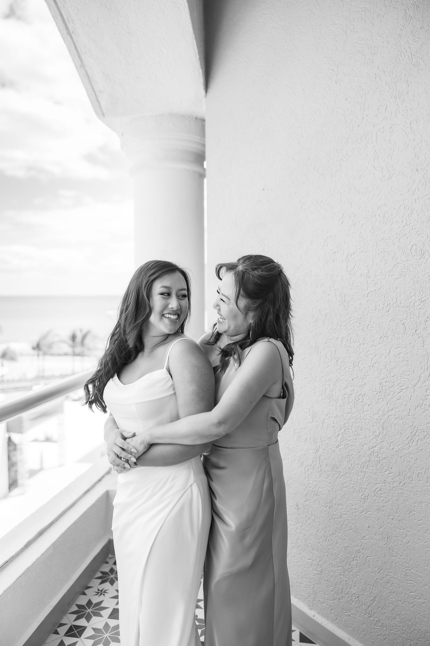 Black and white photo of two women in formal dresses embracing and laughing on a balcony overlooking the ocean.