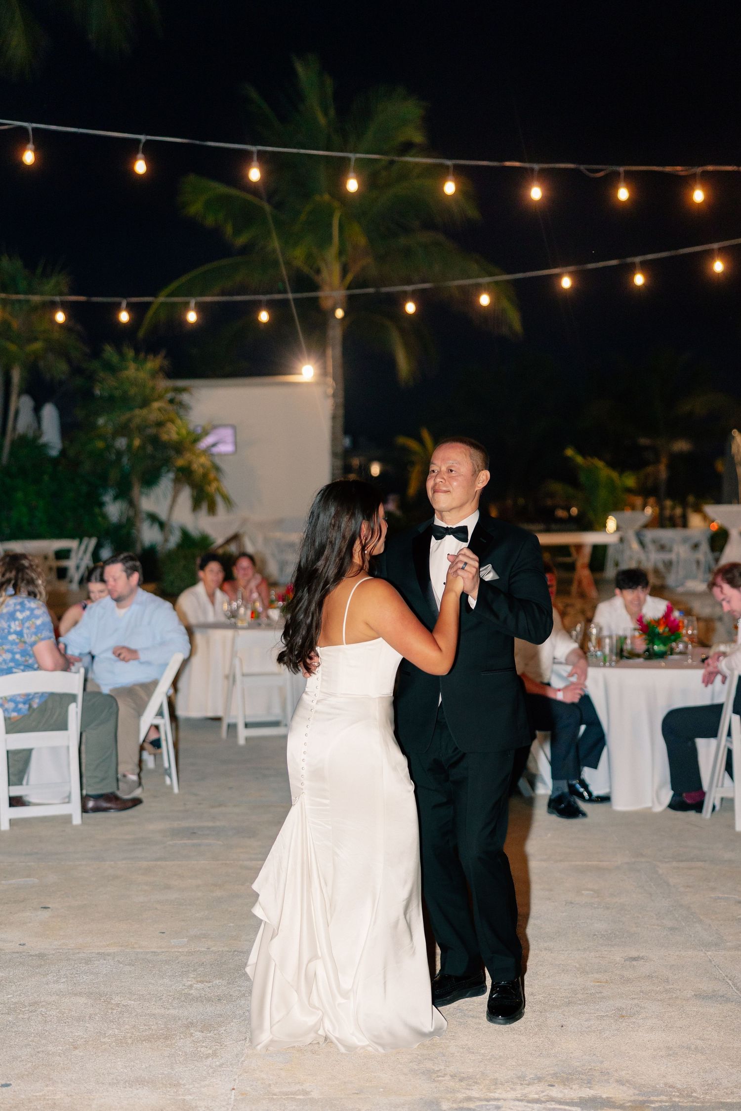 Bride and groom share their first dance under string lights at an outdoor tropical wedding reception at night.