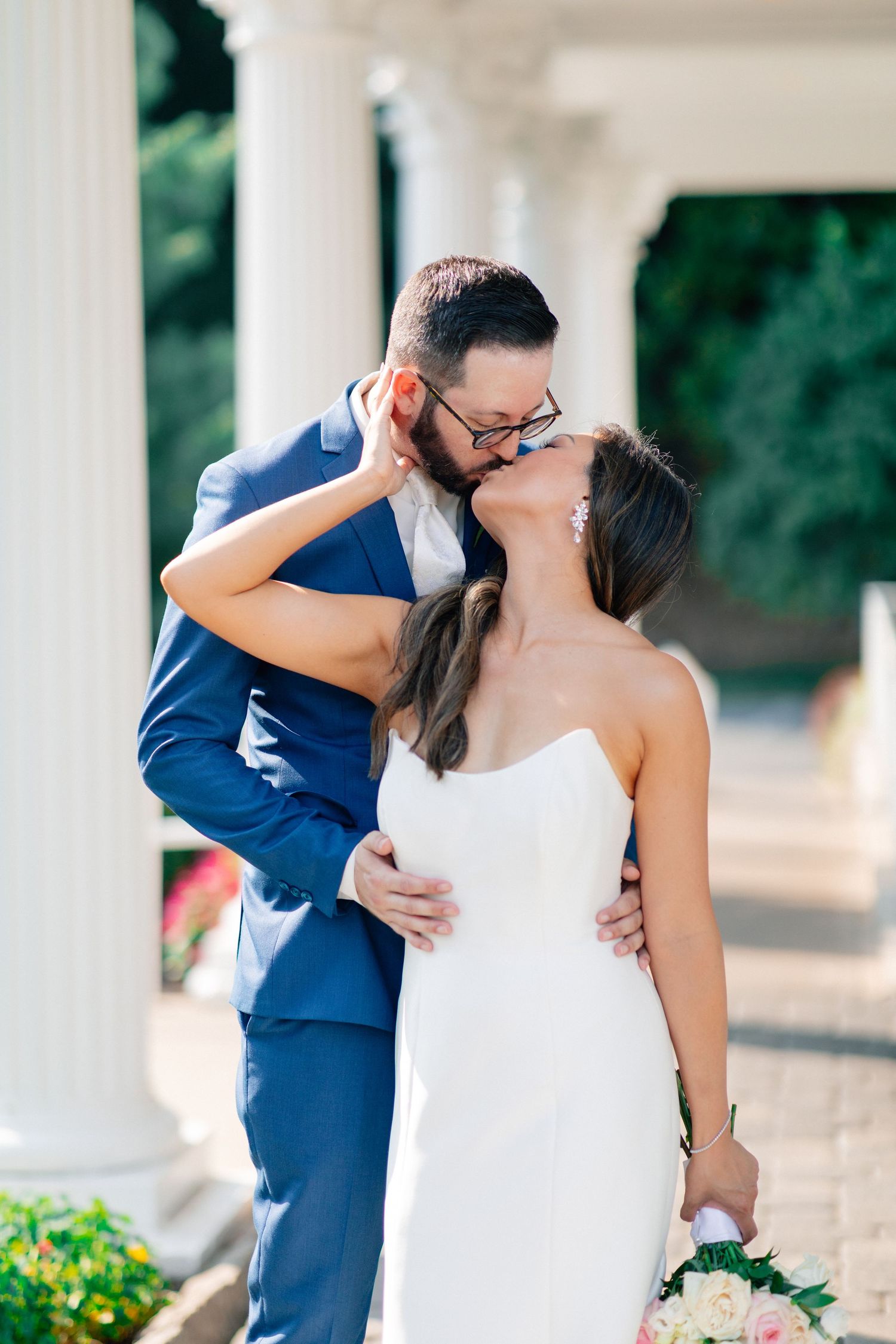 A bride and groom share a romantic kiss outdoors, with white columns in the background, as she holds a bouquet.