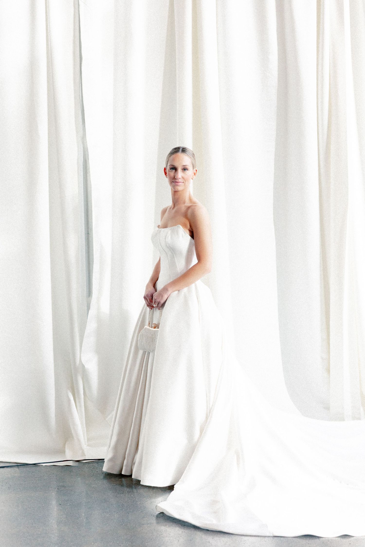 Elegant bride in a strapless white satin ball gown with a flowing train, posed before white draped curtains.