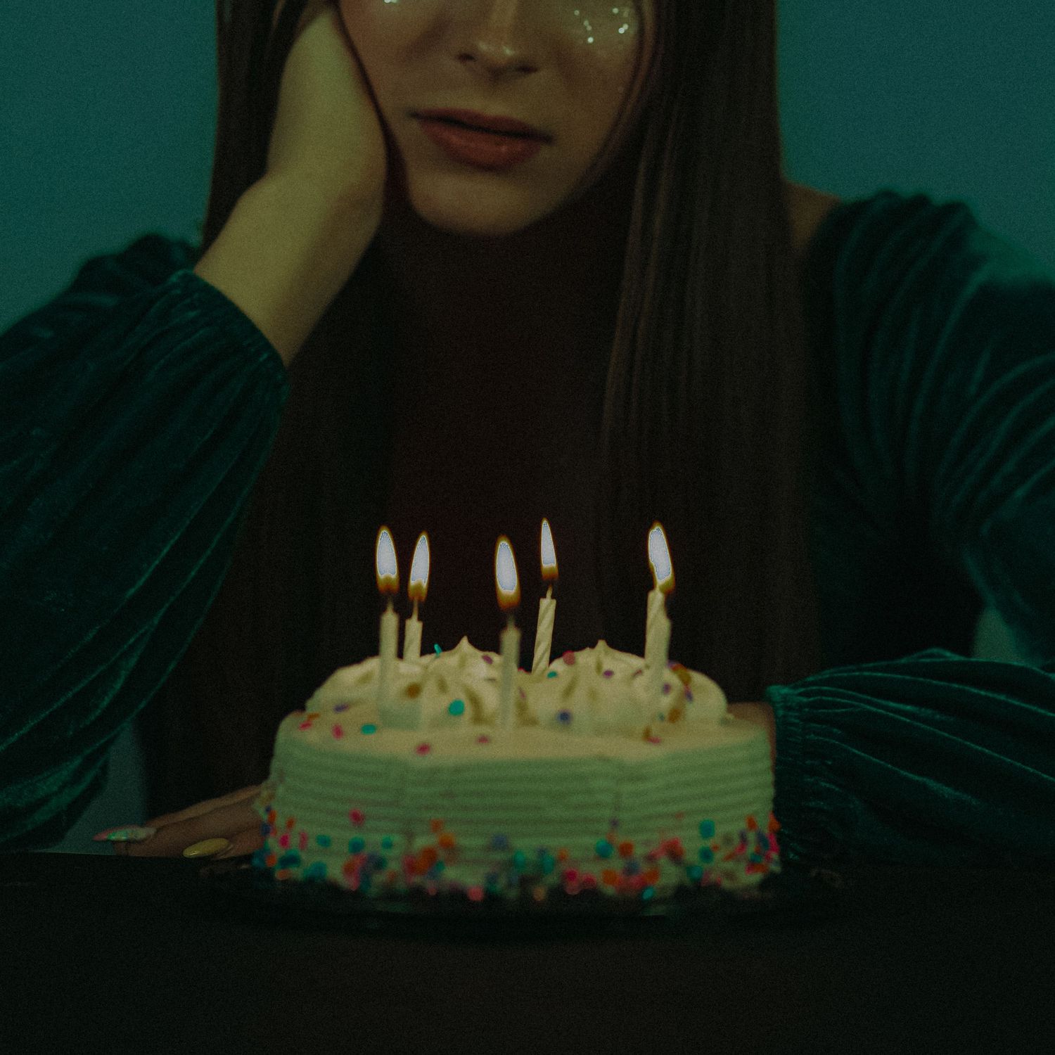 A melancholy woman with glitter eyeshadow rests her head on her hand beside a birthday cake with lit candles.