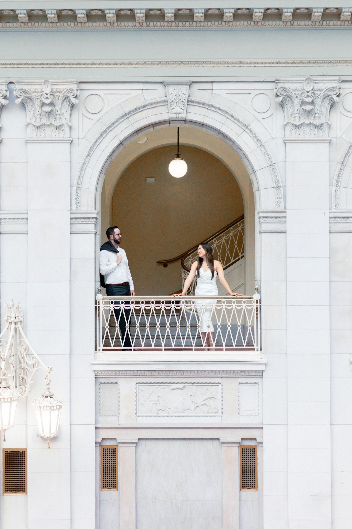 Bride and groom stand on an ornate balcony framed by a stunning white stone archway with classical details.