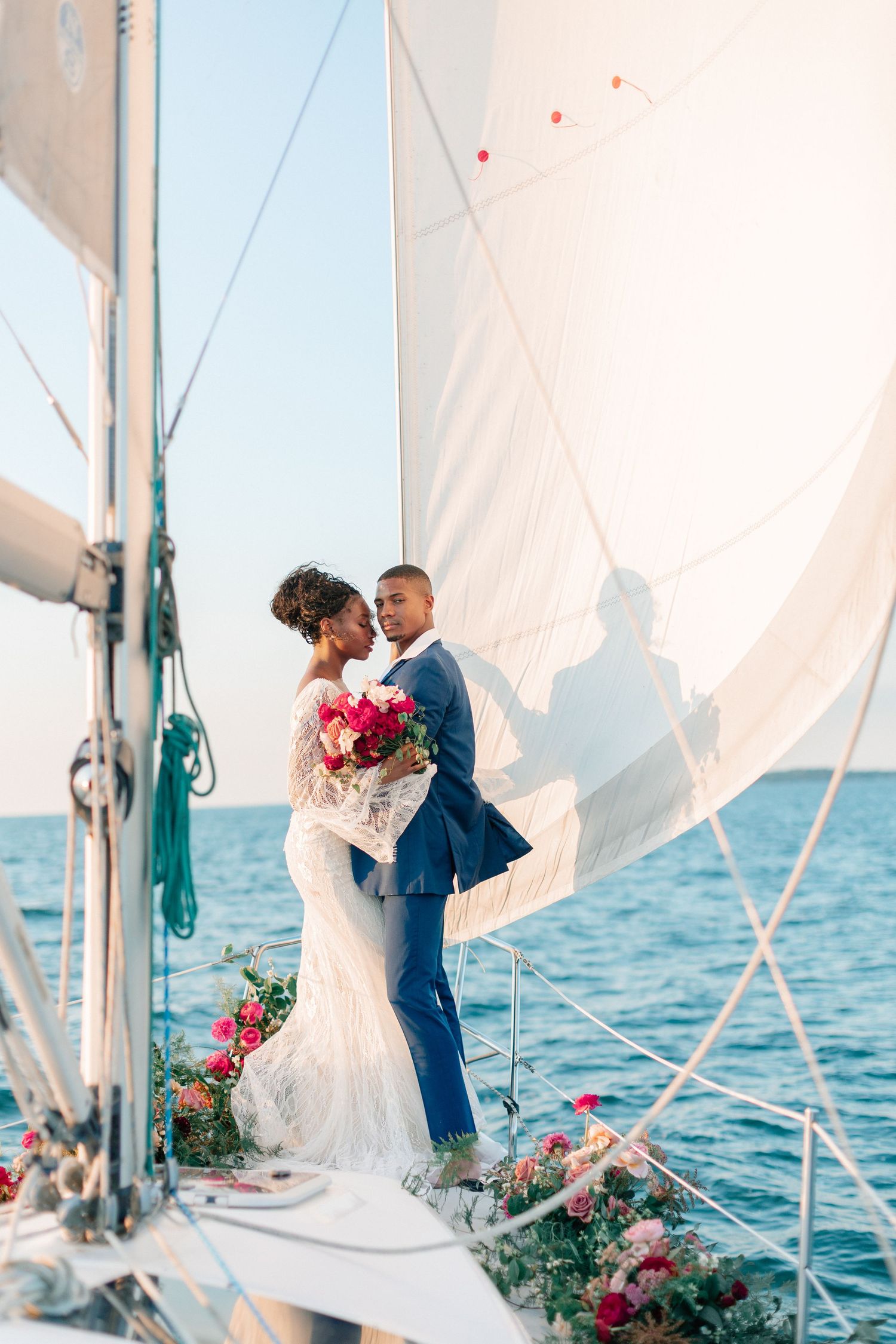 Bride and groom embracing on a sailboat deck with white sails, ocean backdrop, and colorful floral arrangements.