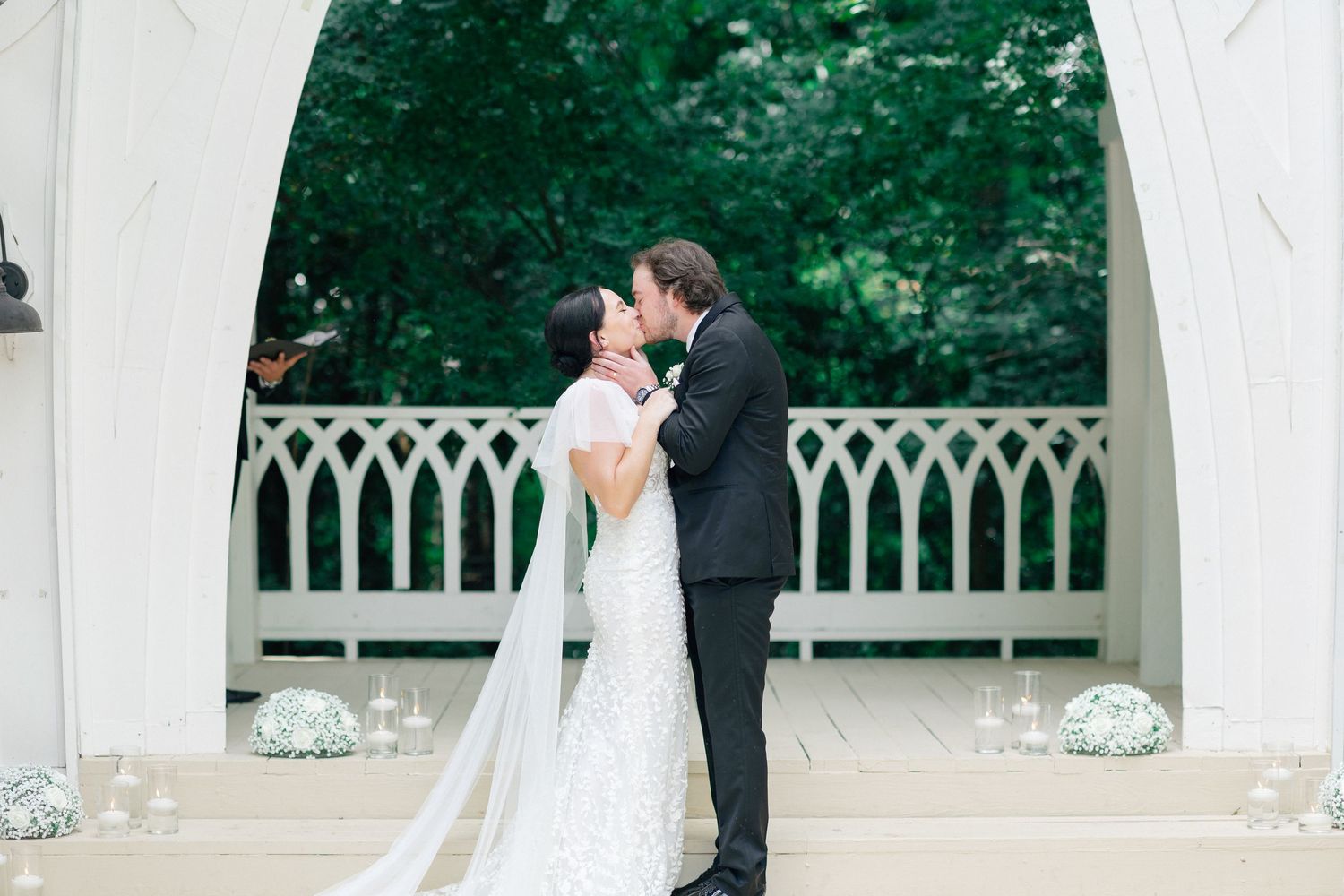 Bride and groom share first kiss at elegant white gazebo wedding ceremony surrounded by lush greenery.