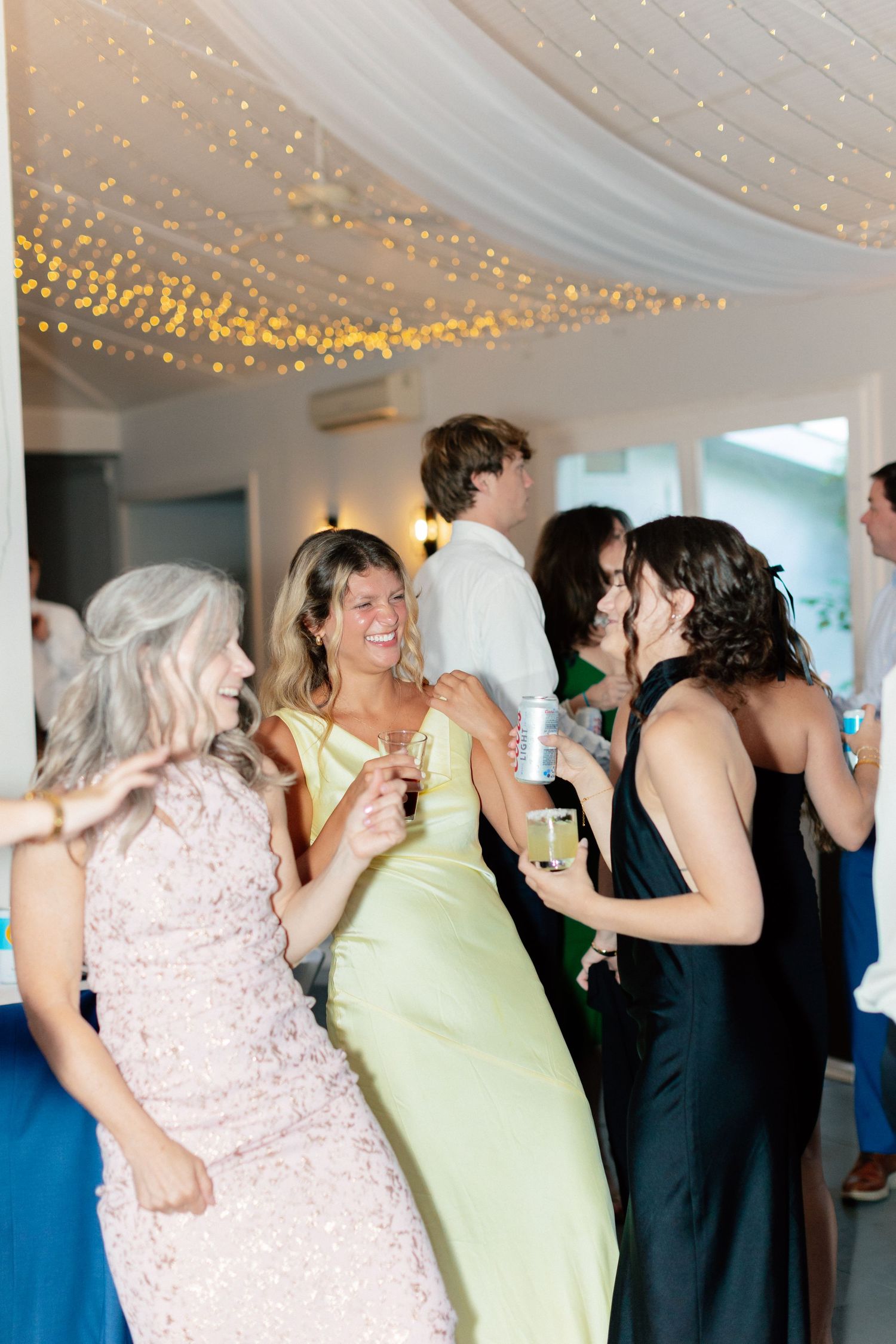 Guests in elegant dresses laugh and mingle at a wedding reception under twinkling fairy lights.